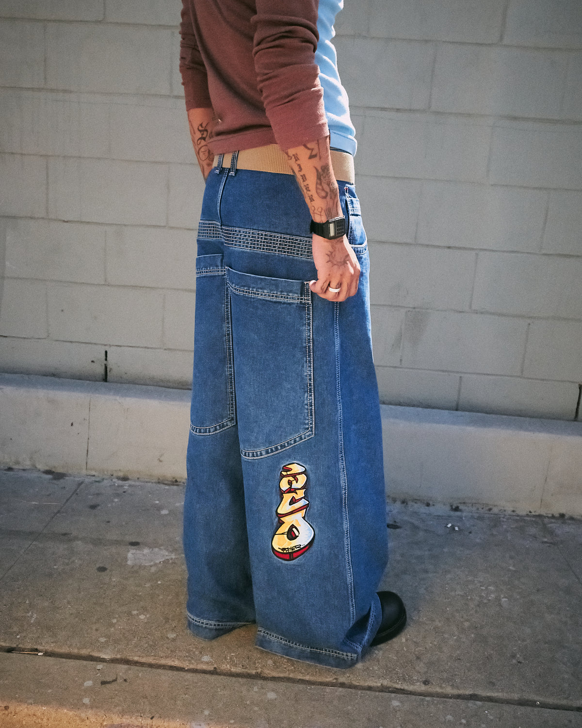Model wearing wide-leg dark stone wash jeans, back-side view with a large embroidered graffiti logo on the leg