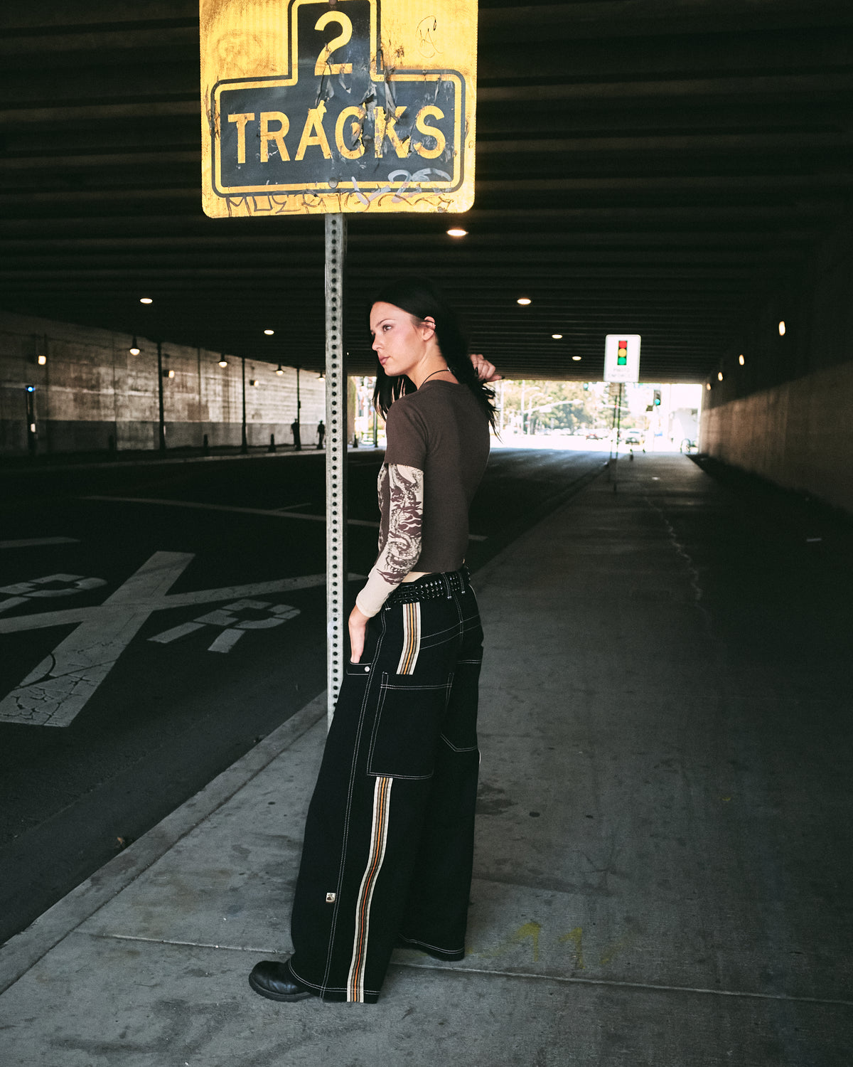 Model standing under a '2 Tracks' street sign in an underpass, wearing wide-leg black jeans with striped tape, side view