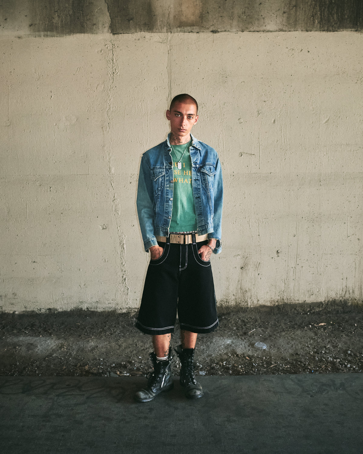 Model wearing the black shorts with a denim jacket, green tee, tan belt and black boots, full-length front view