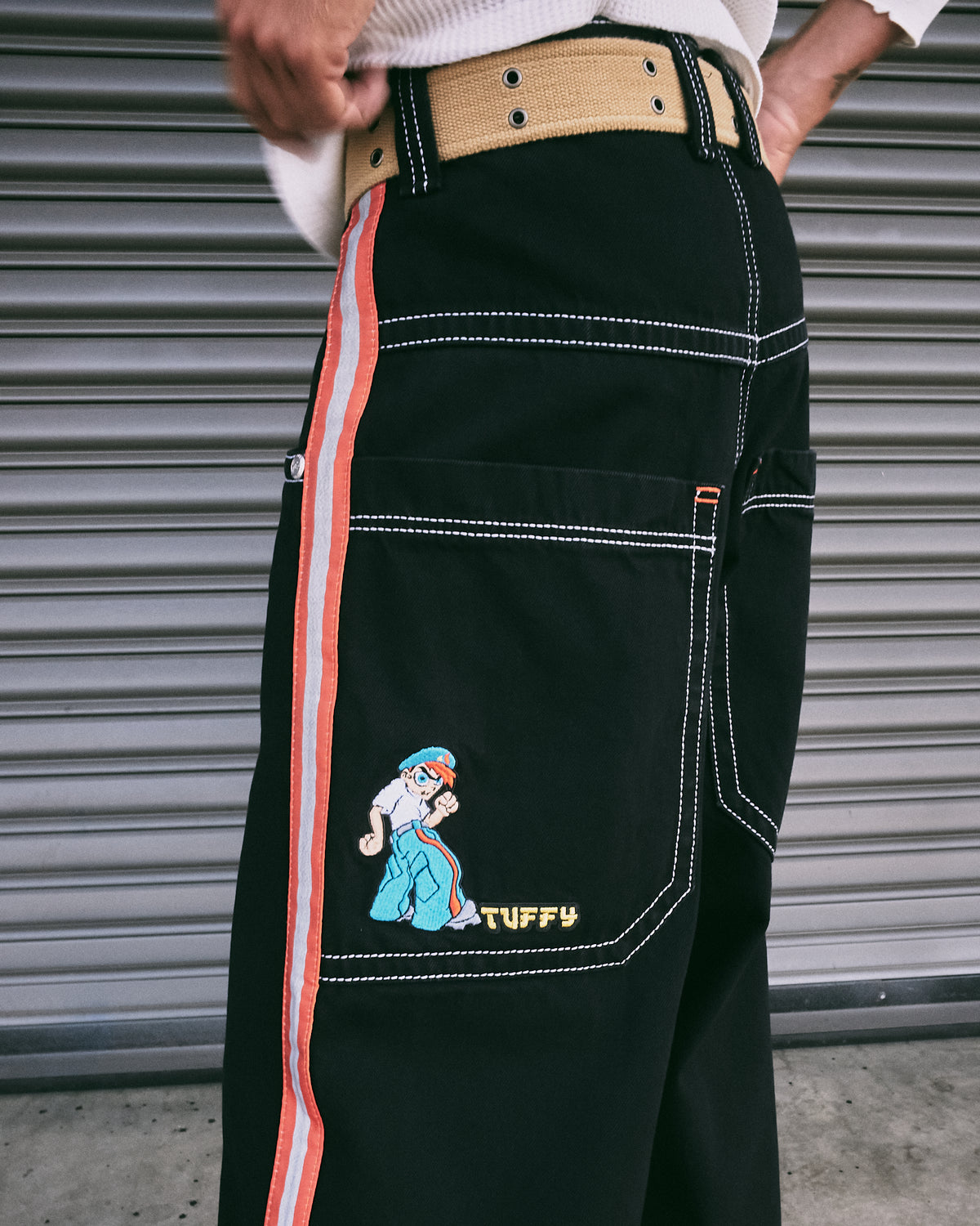 Back side view of the black jeans with a tan canvas belt, white stitching, side stripe and TUFFY cartoon patch on the pocket
