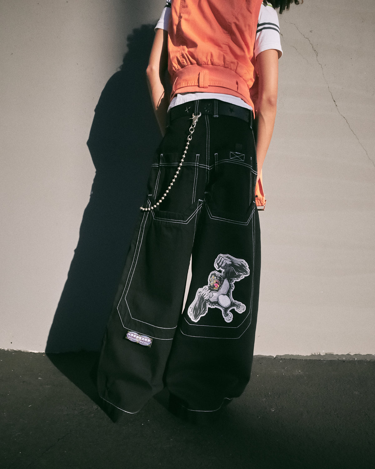 Back view of a model wearing wide-leg black jeans with a gorilla patch on one back pocket and a wallet chain