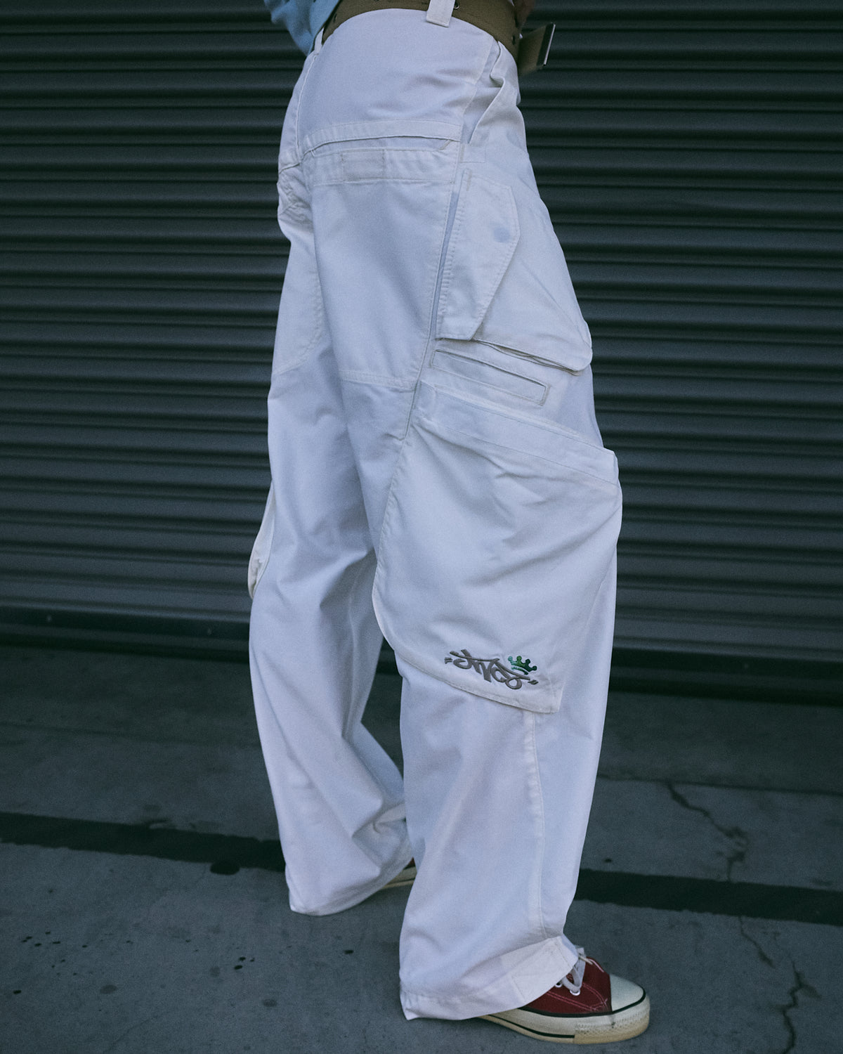 Side view of off-white wide-leg cargo pants on a model, showing layered cargo pockets with embroidered JNCO logo