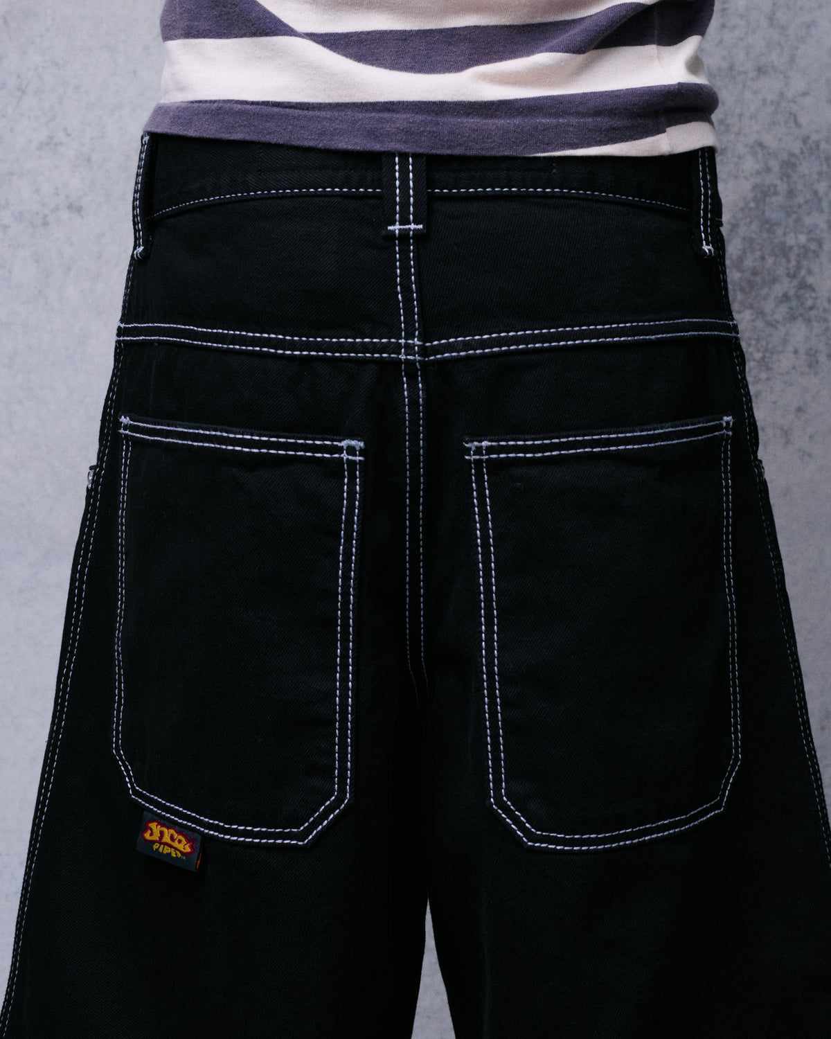 Back view of a model wearing the black denim shorts, showing both back pockets, white stitching and JNCO Pipes tab
