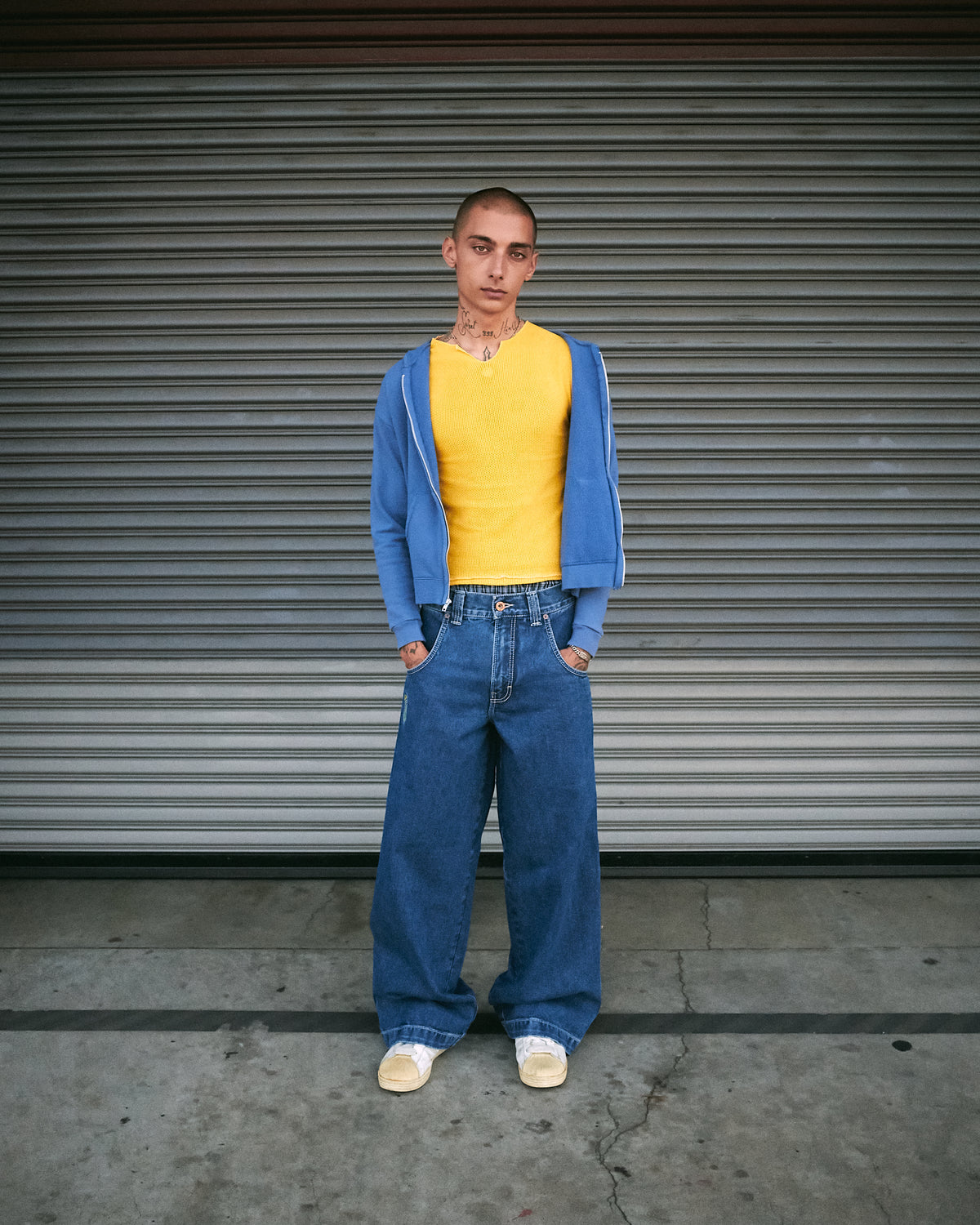 Model standing front-on in dark stone wash wide-leg jeans with a yellow top and blue zip hoodie, hands in pockets