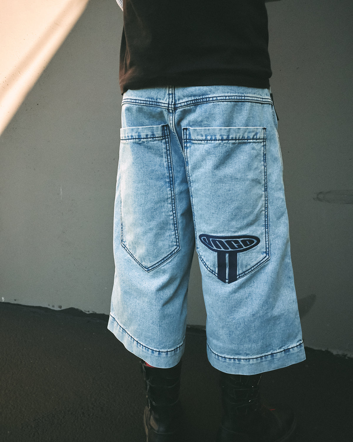 Back view of light blue wide denim shorts worn outdoors, showing long back pockets and navy JNCO embroidery