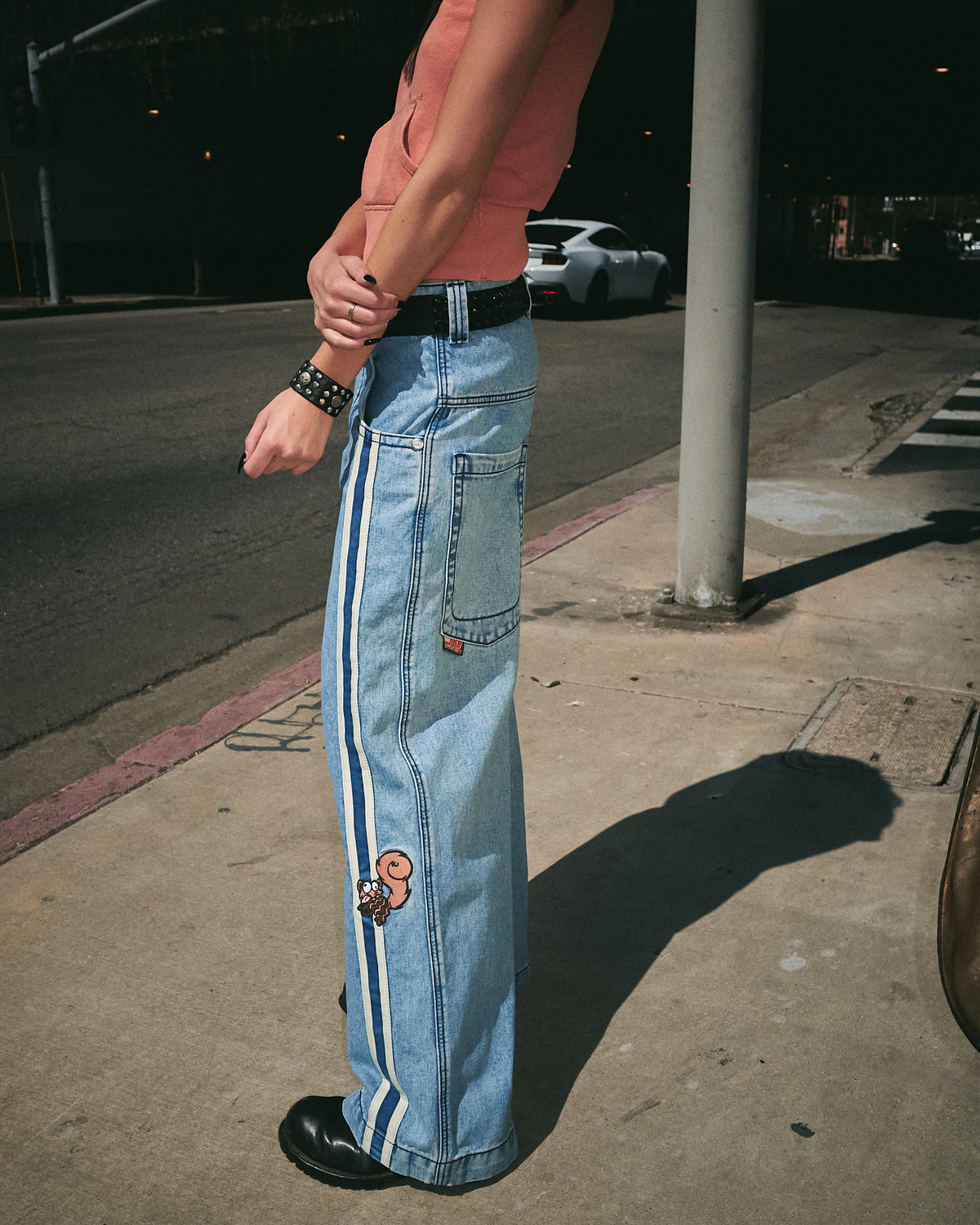 Model on a sidewalk wearing the light-wash wide-leg jeans with a black belt, side view showing leg tape and back pocket