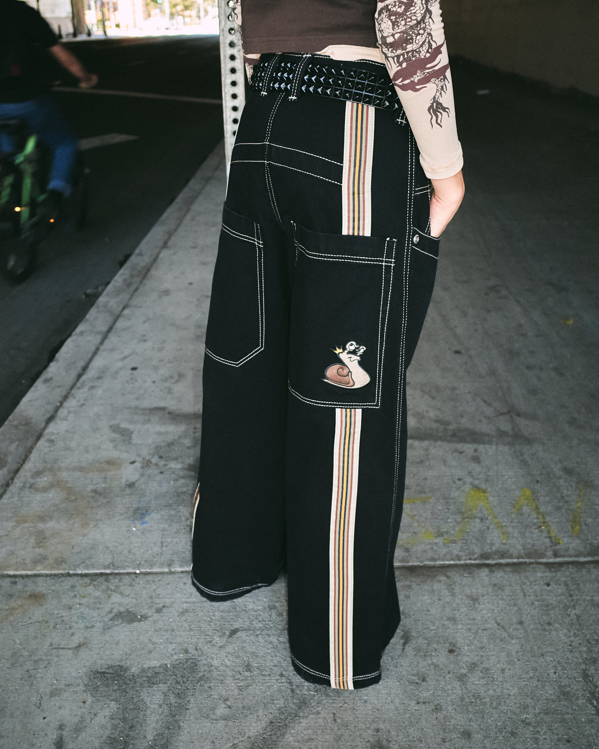 Back view of a model wearing wide-leg black jeans with striped tape down the legs and a snail patch on the pocket