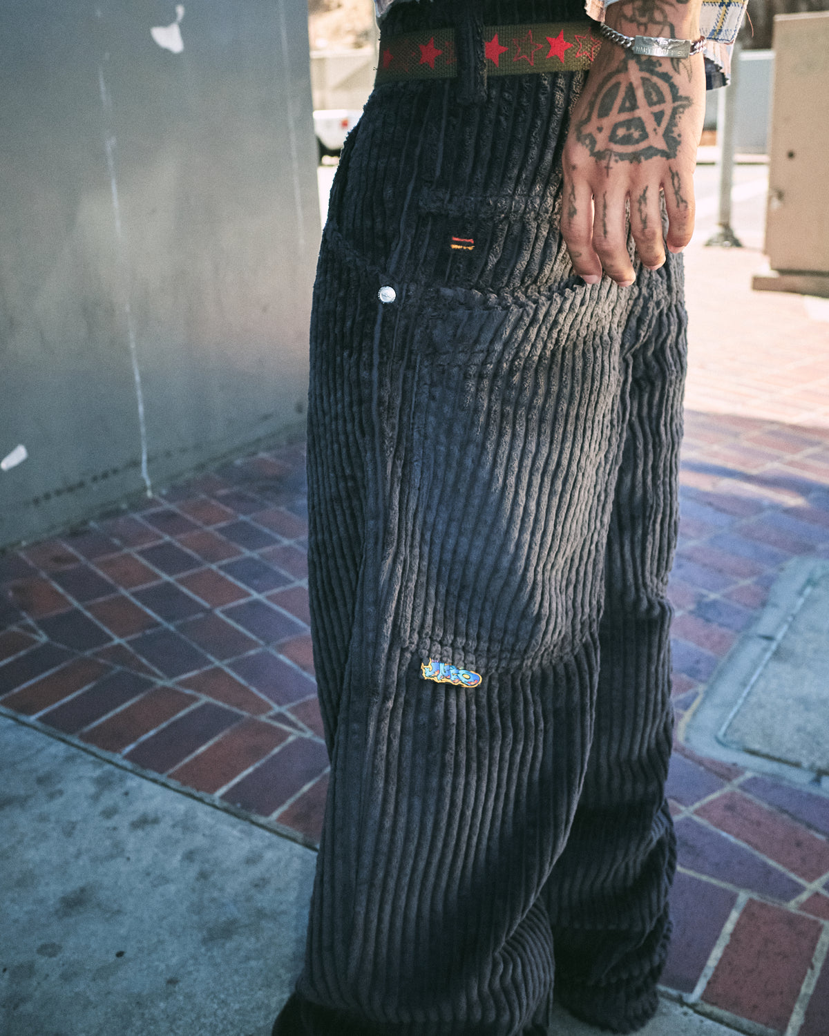 Model's hand at the pocket of dark grey wide-leg corduroy pants with a small embroidered logo, side view outdoors