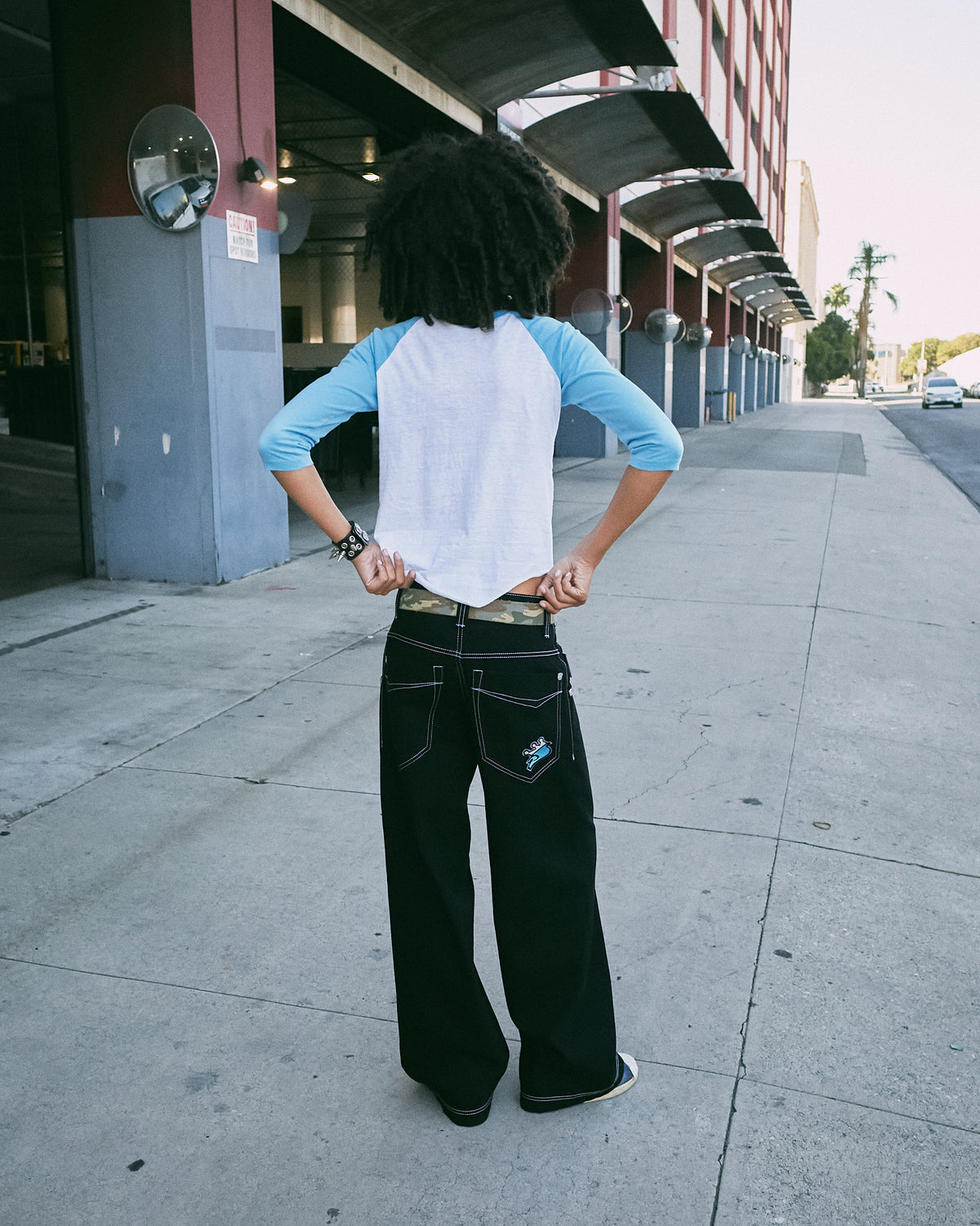 Model on a sidewalk seen from behind wearing black wide-leg jeans with a camo belt and a raglan tee