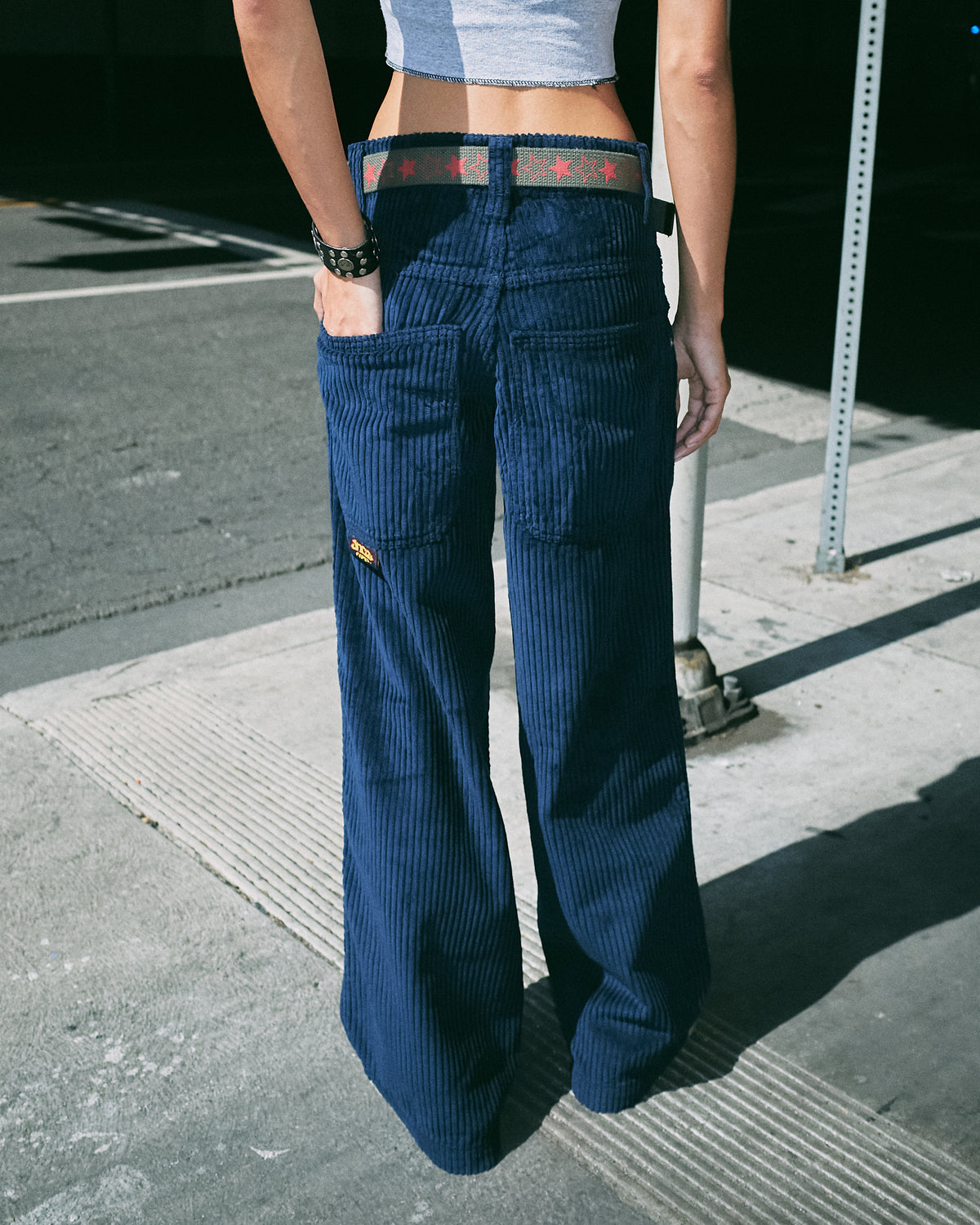 Model on a street corner wearing navy wide-leg corduroy pants with a star belt, full back view
