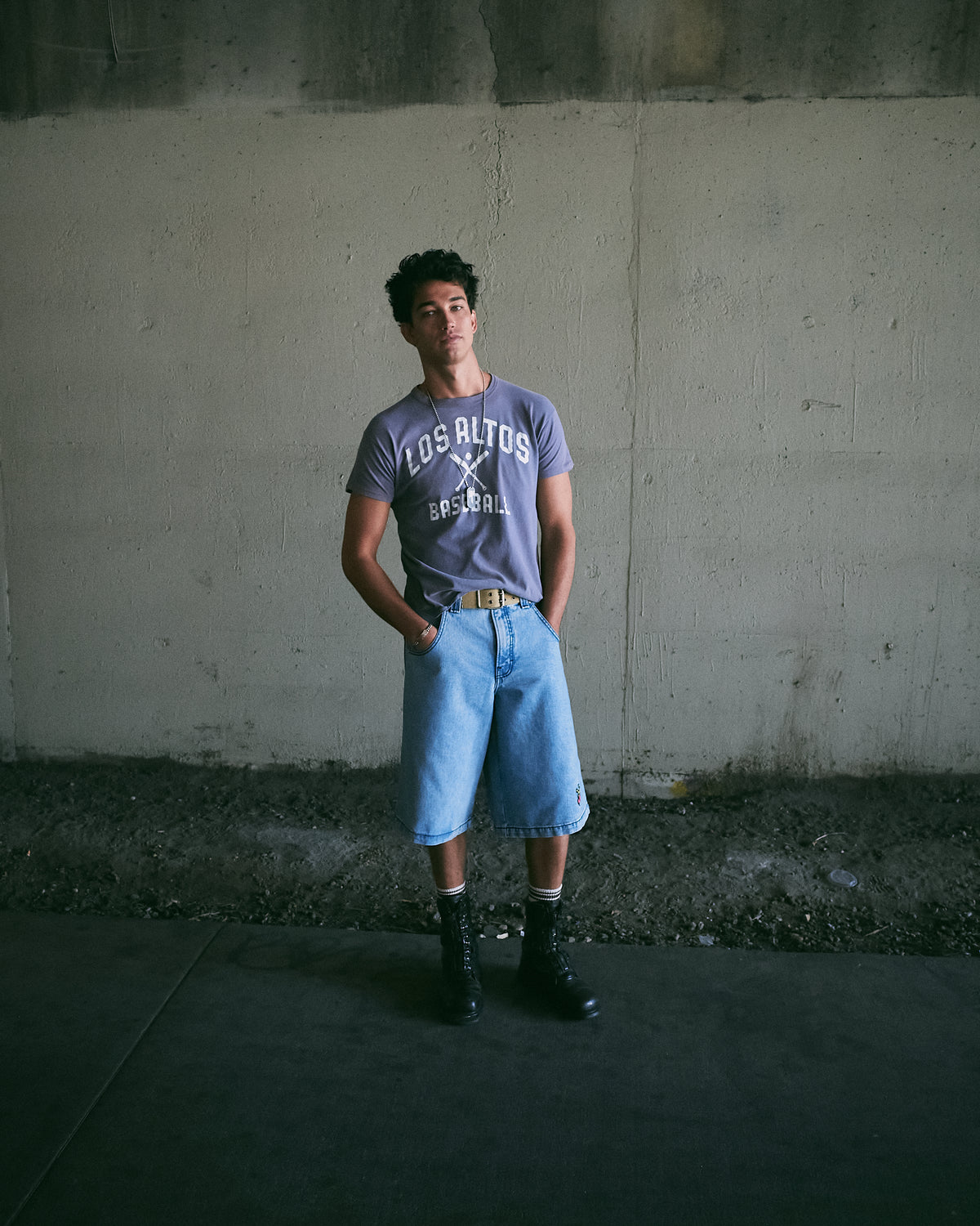 Model standing against a concrete wall wearing light blue knee-length denim shorts, a tan belt and black boots