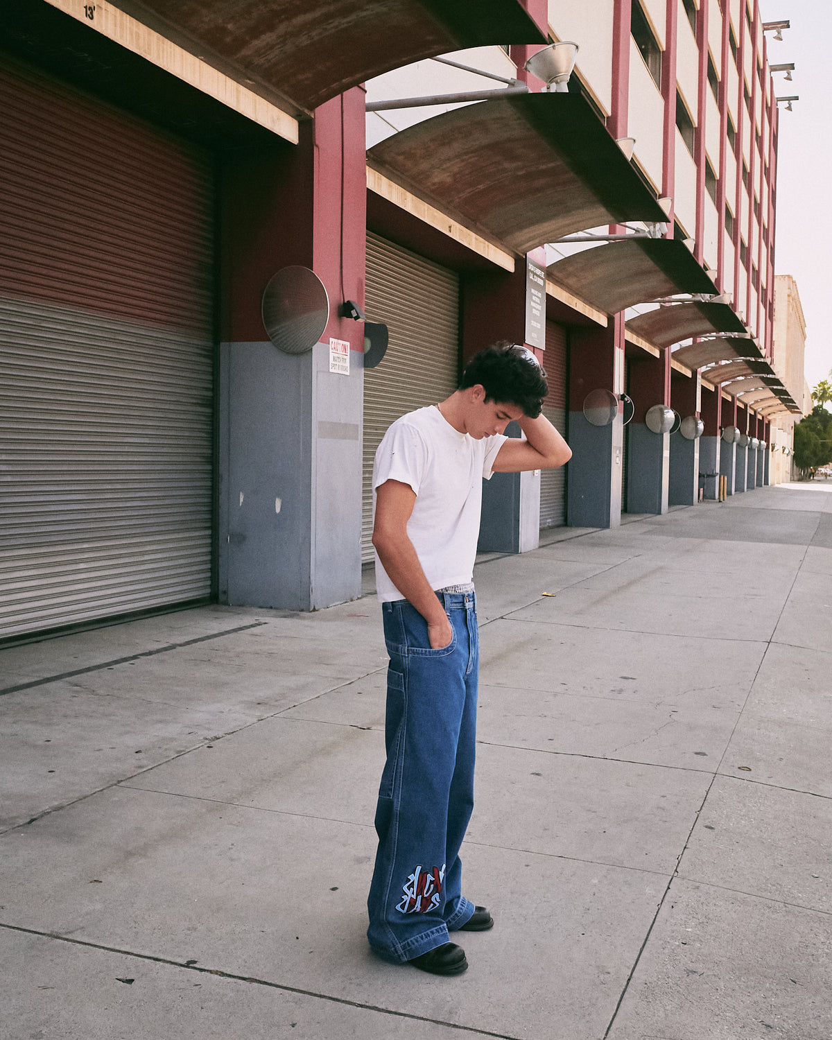 Model in a white T-shirt wearing wide-leg dark stone wash jeans with graffiti embroidery near the hem, on a sidewalk
