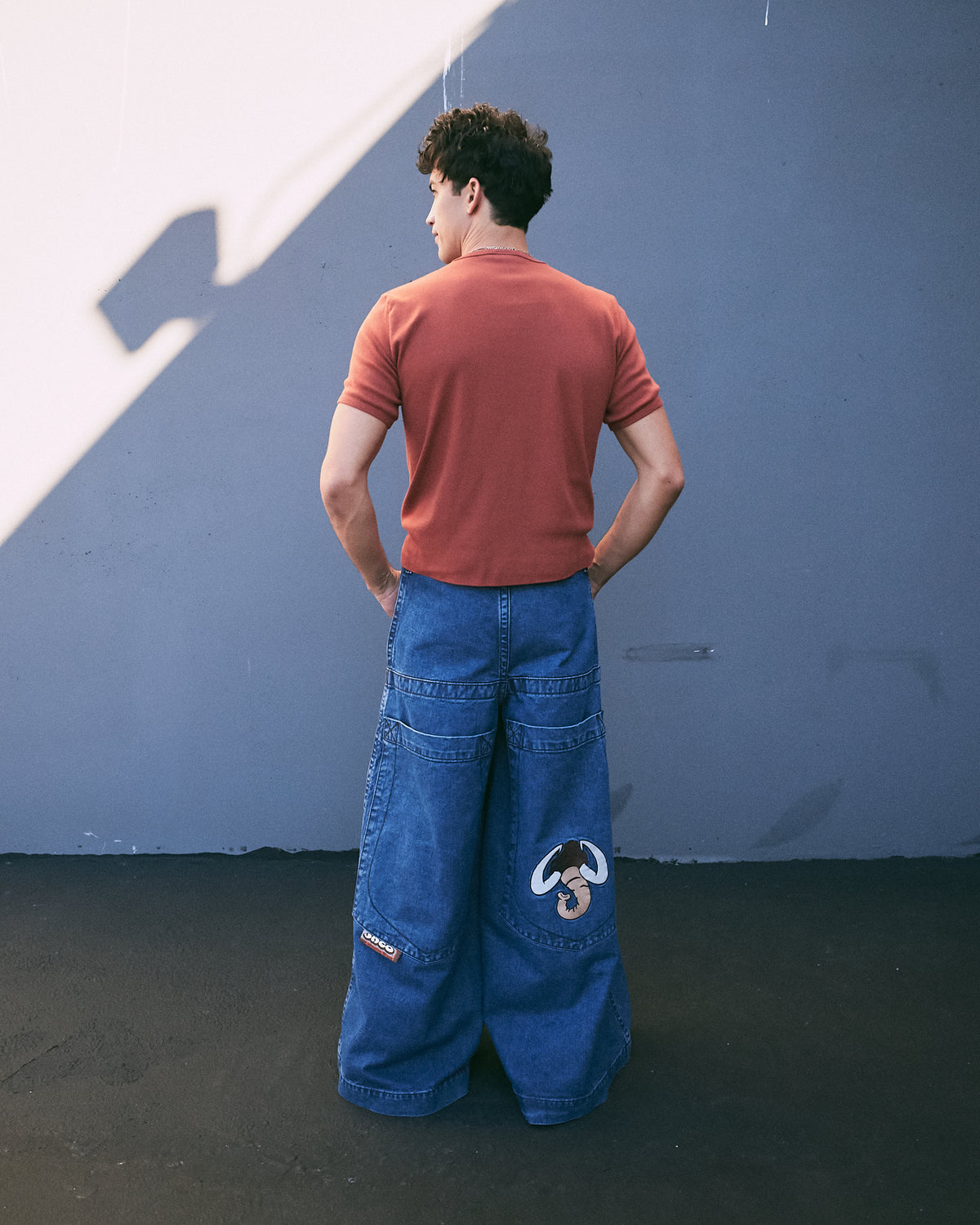 Model in a rust T-shirt wearing extra-wide dark stone wash jeans, back view against a grey wall