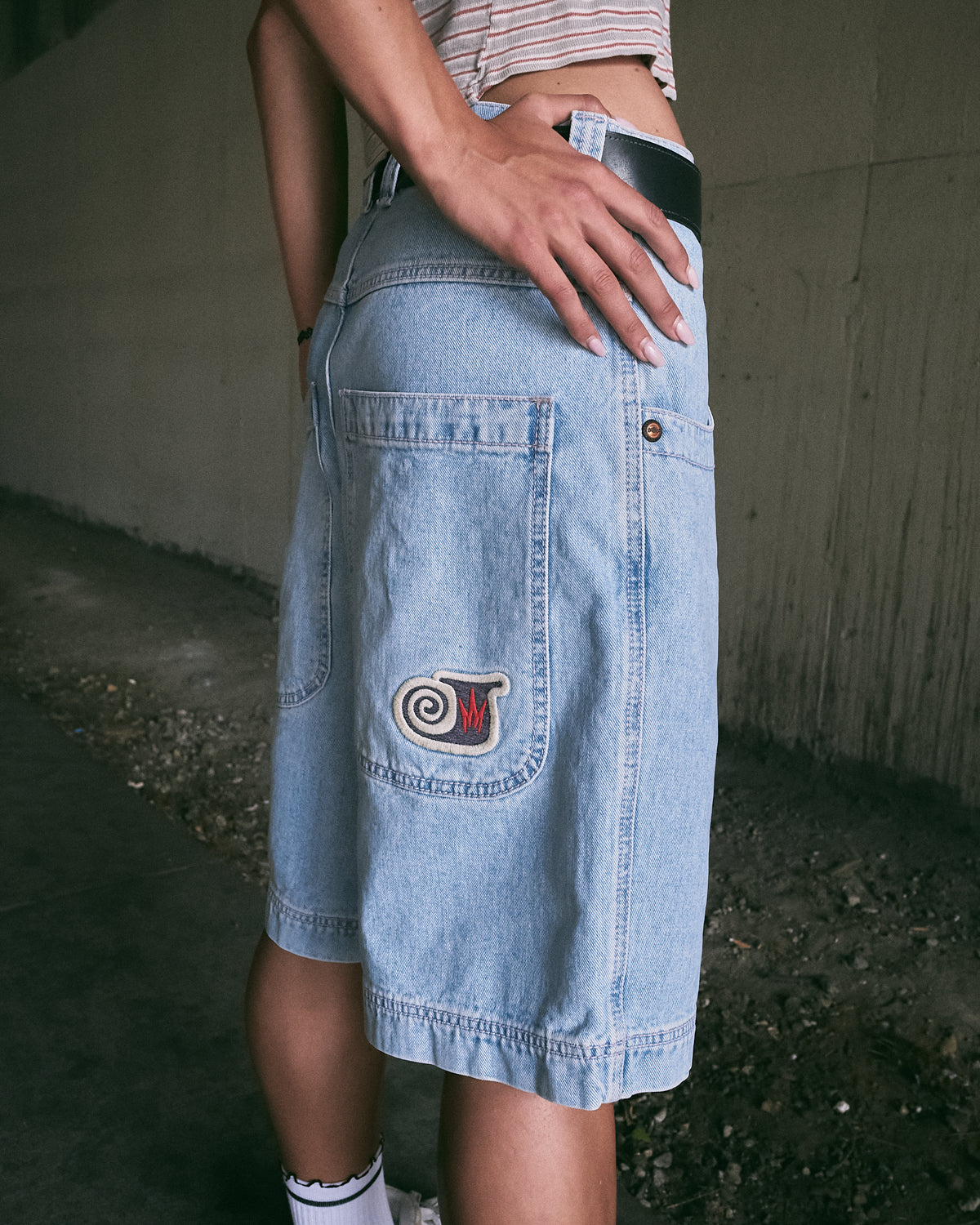 Side-back view of the model in light-wash denim shorts, hand on hip, embroidered swirl patch on the back pocket