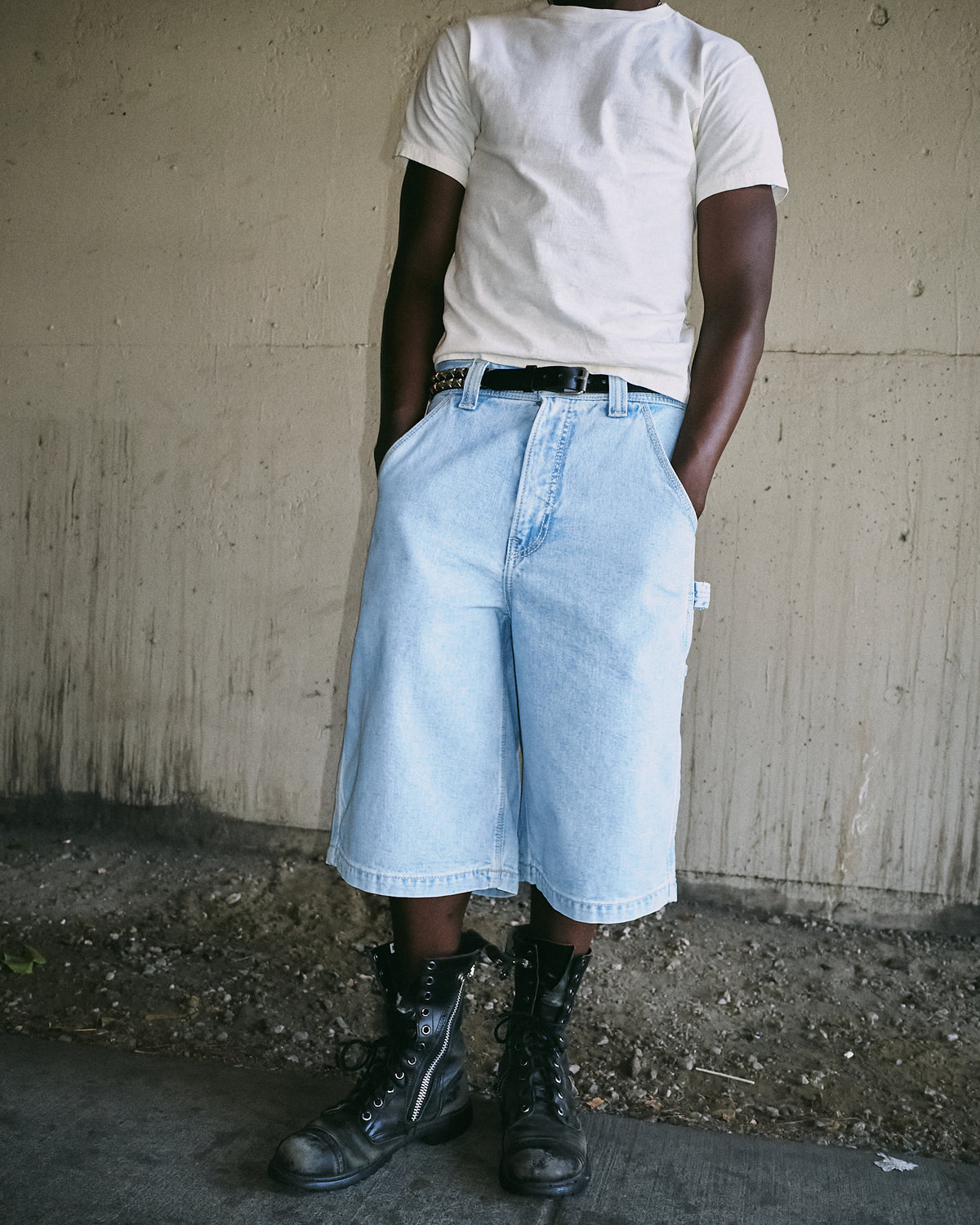 Model wearing light-wash knee-length carpenter shorts with a white T-shirt, studded belt and black boots, front view