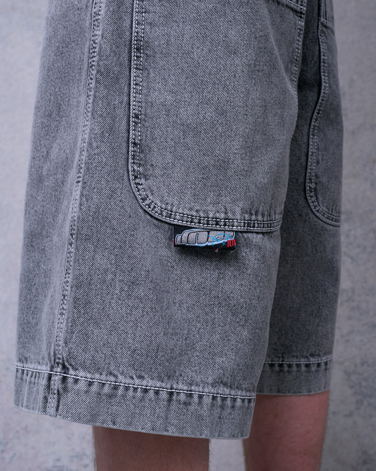 Close-up of faded black denim shorts on a model, showing the side pocket, small JNCO tab label and stitched hem