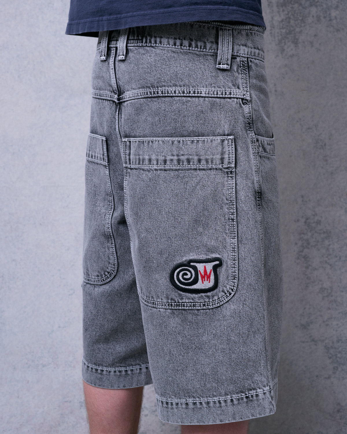 Model wearing faded black denim shorts, rear three-quarter view of the back pocket with swirl J embroidery