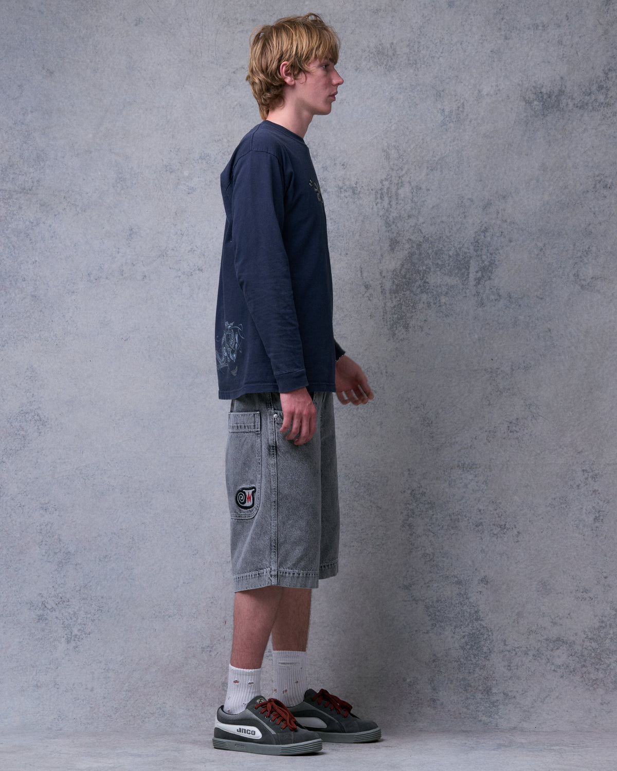 Model wearing washed black denim shorts, side view showing the side pocket with an embroidered patch