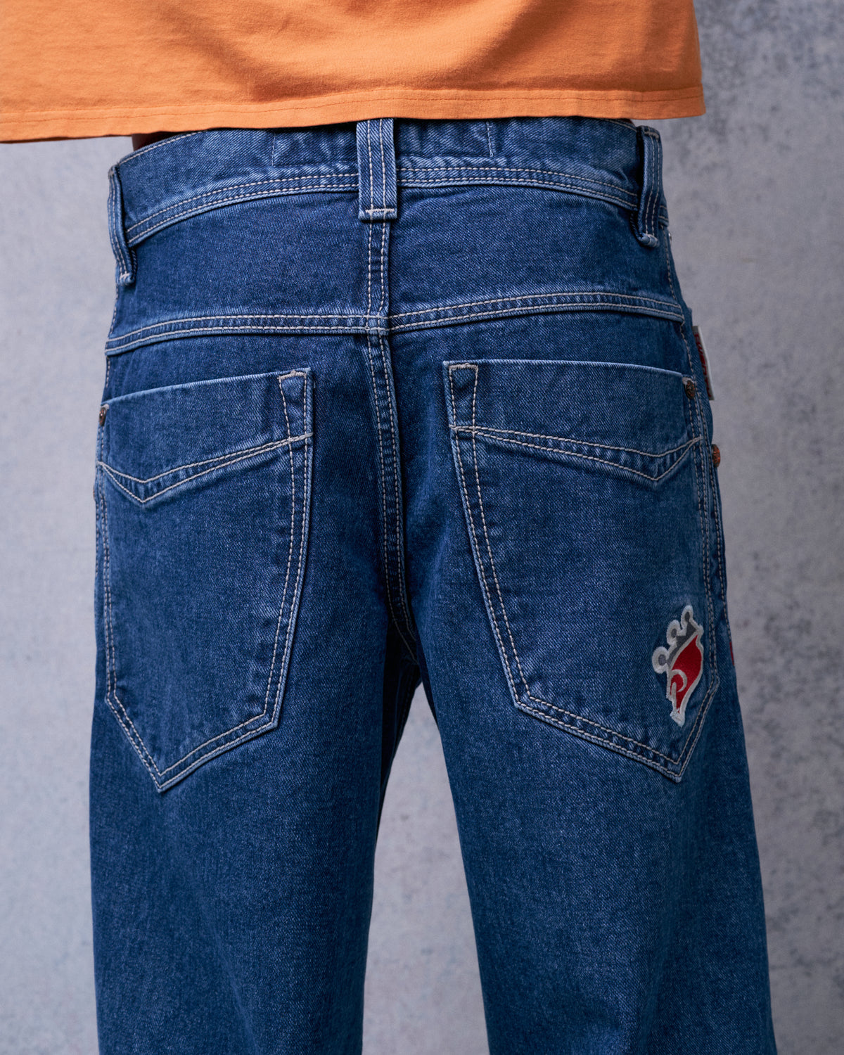 Back view of dark stone wash jeans on a model, two back pockets with a red crown embroidery on one
