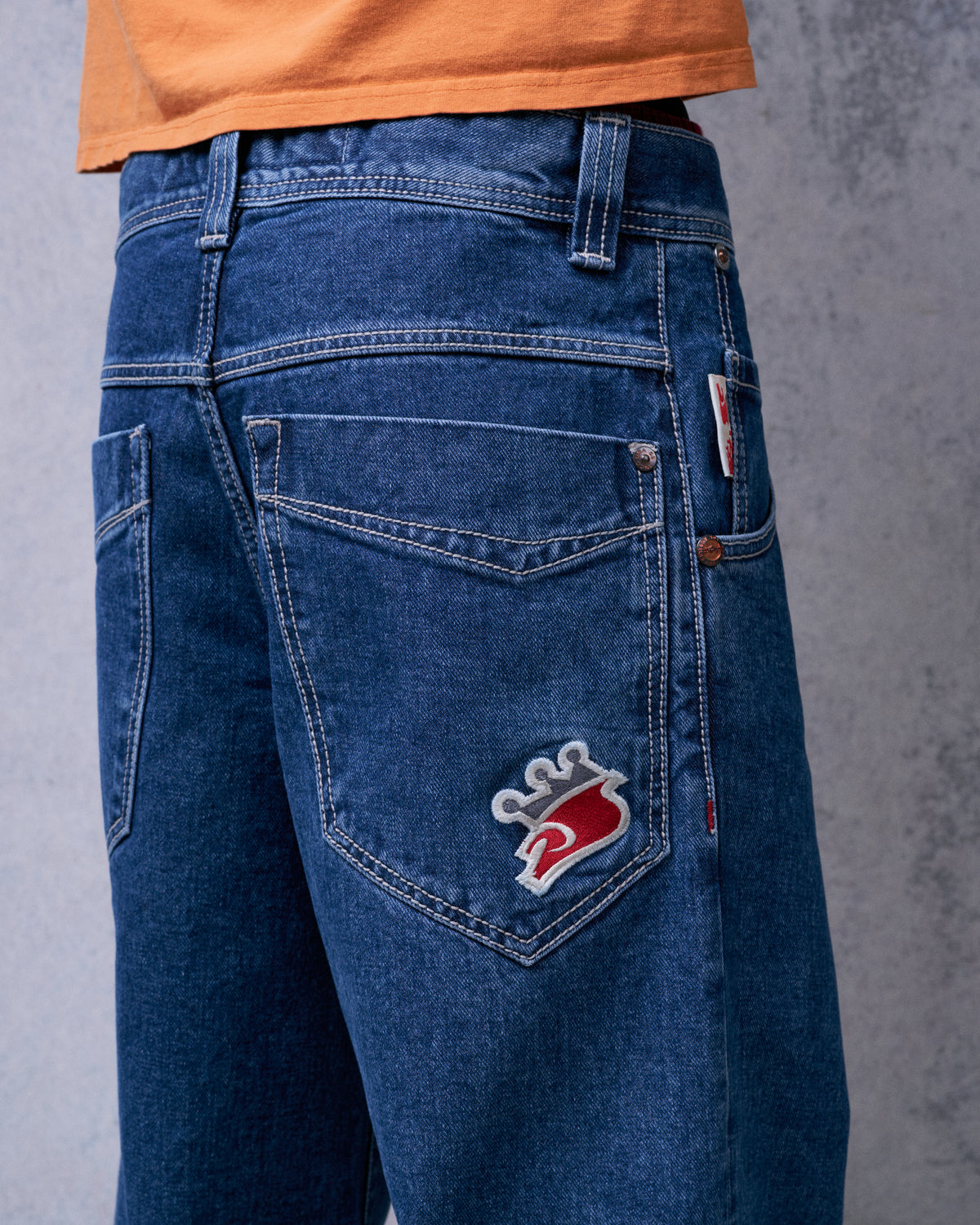 Angled back view of dark stone wash jeans showing the red and grey crown embroidery on the back pocket