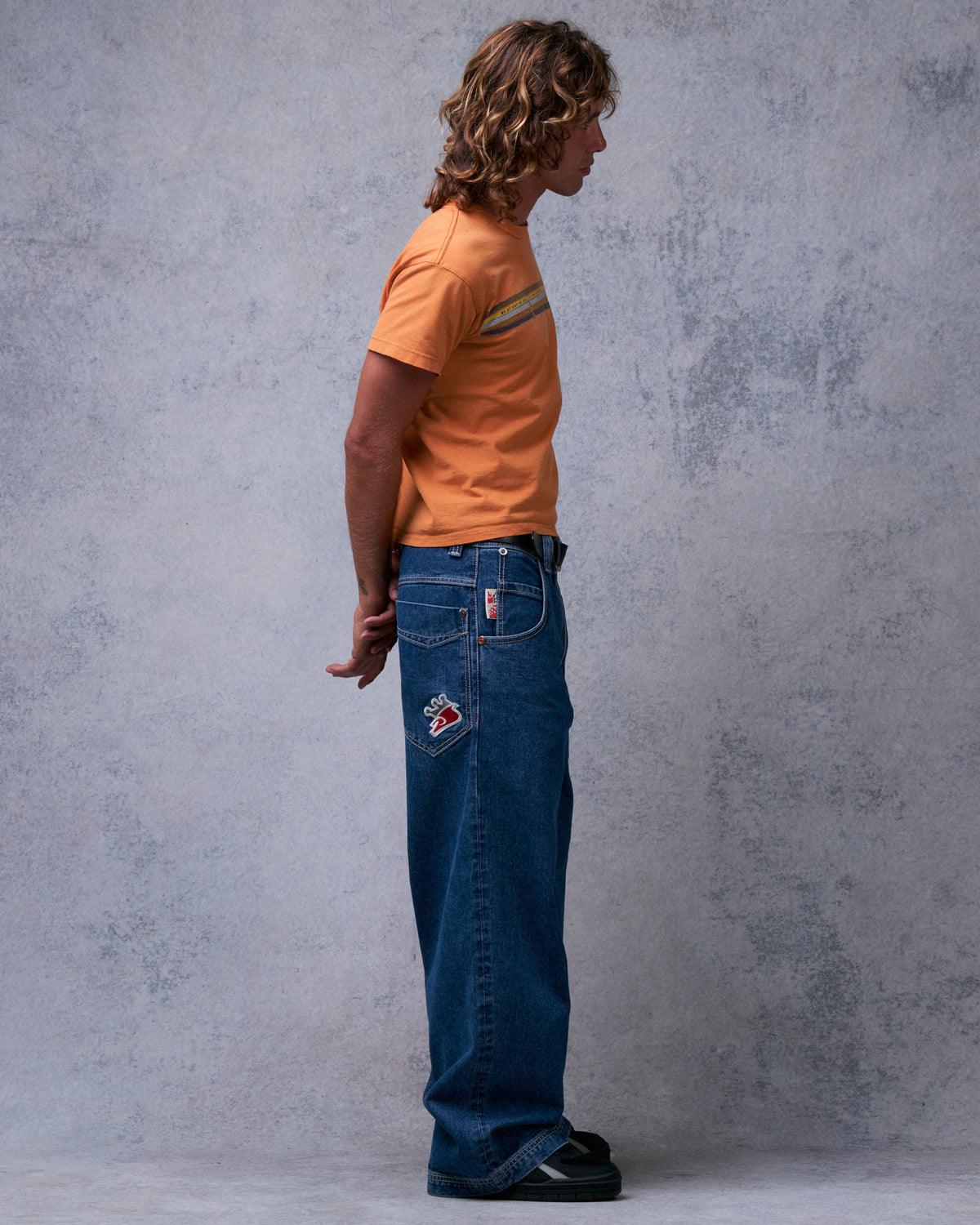 Model wearing dark stone wash wide-leg jeans, side view showing the crown patch on the back pocket and red side tab