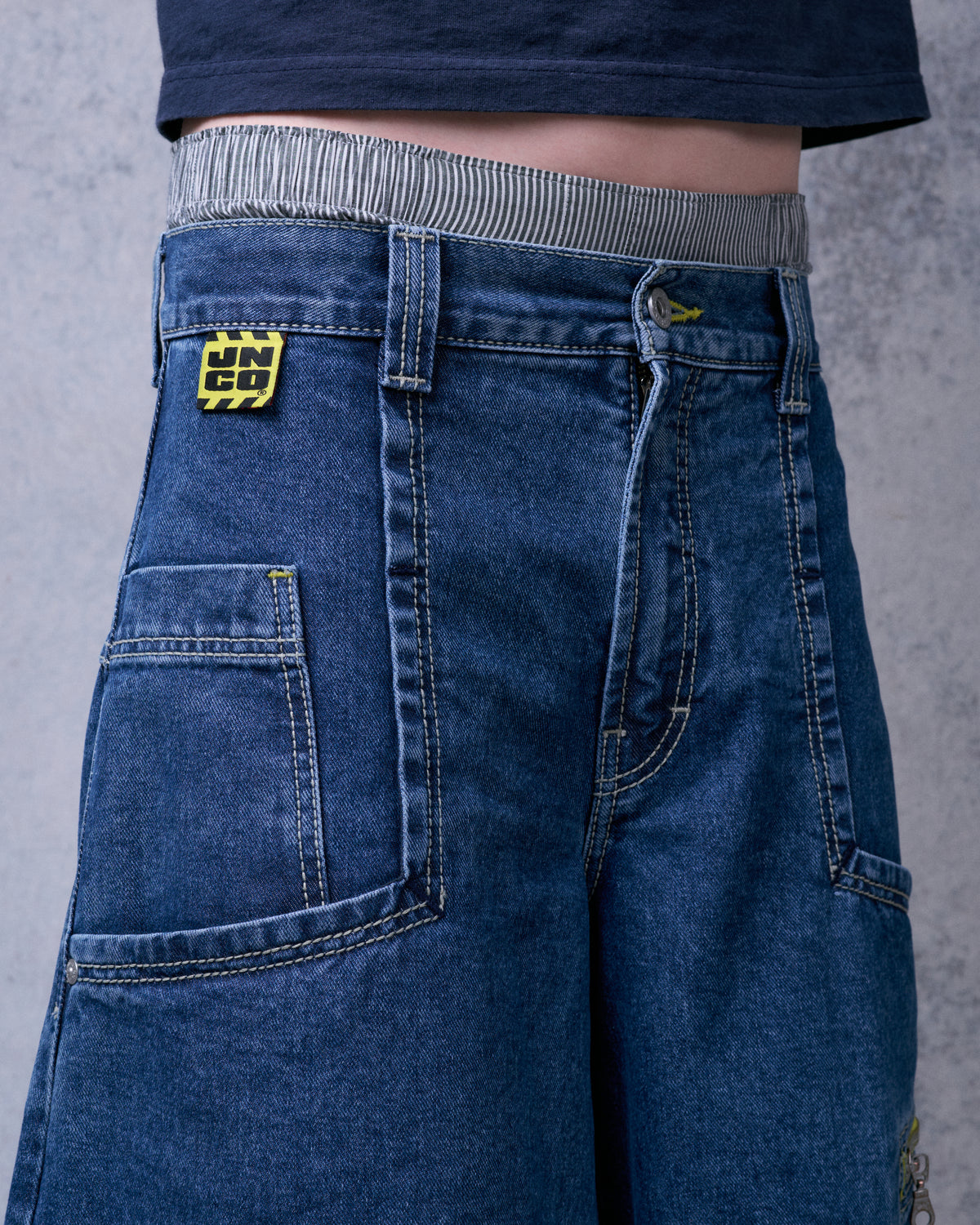Model wearing the jeans, front waist close-up with yellow JNCO patch, button fly and front patch pocket