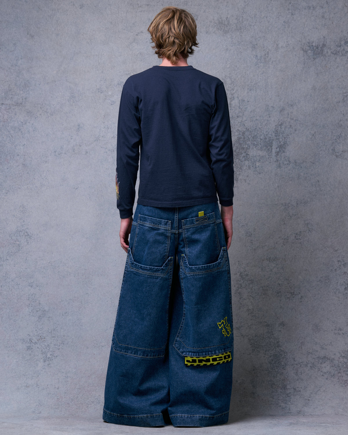 Model wearing the dark stone jeans, straight back view showing long back pockets, yellow Crime Scene embroidery and JNCO tag