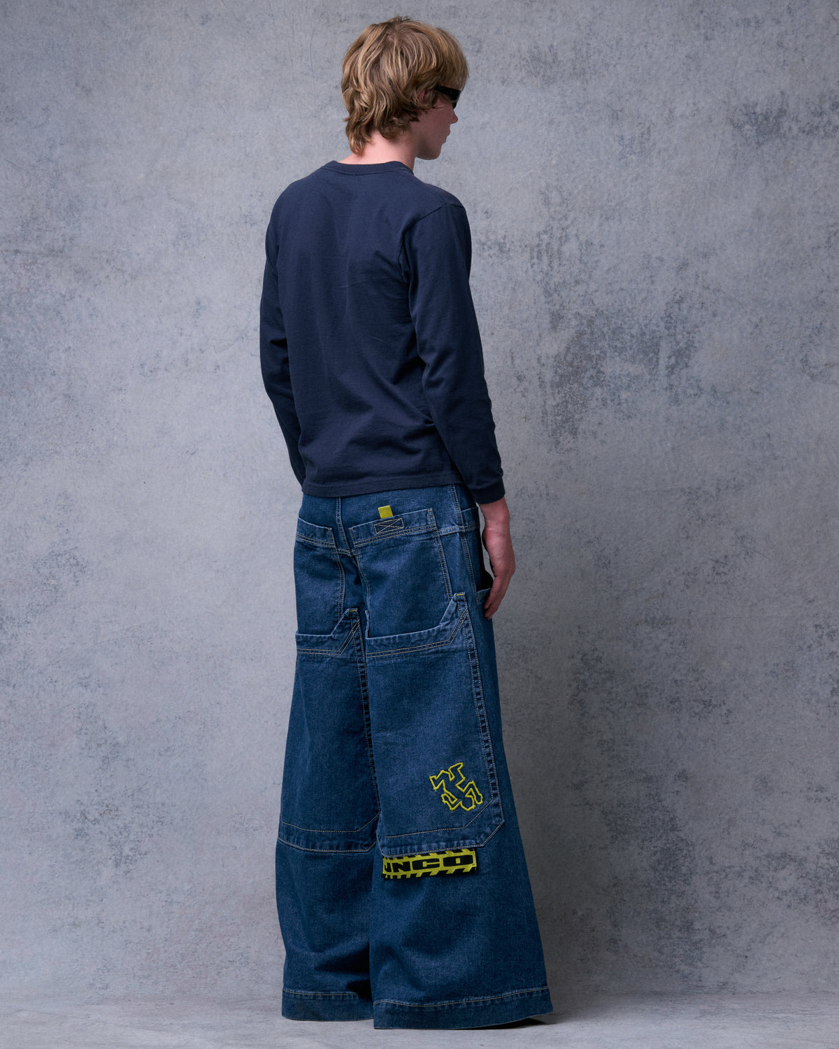 Model wearing the dark stone jeans, back three-quarter view of oversized back pockets with yellow embroidery and JNCO tag