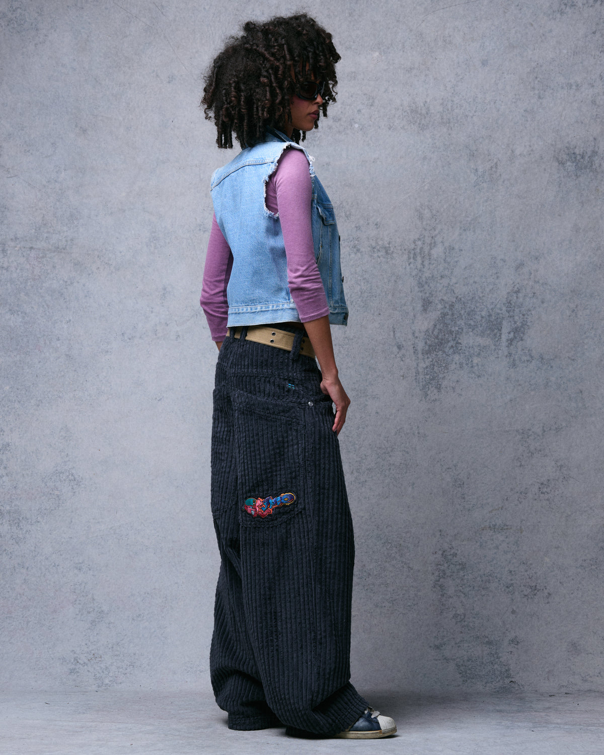 Model wearing dark charcoal wide-leg corduroy pants, side-back view showing the embroidered back pocket patch
