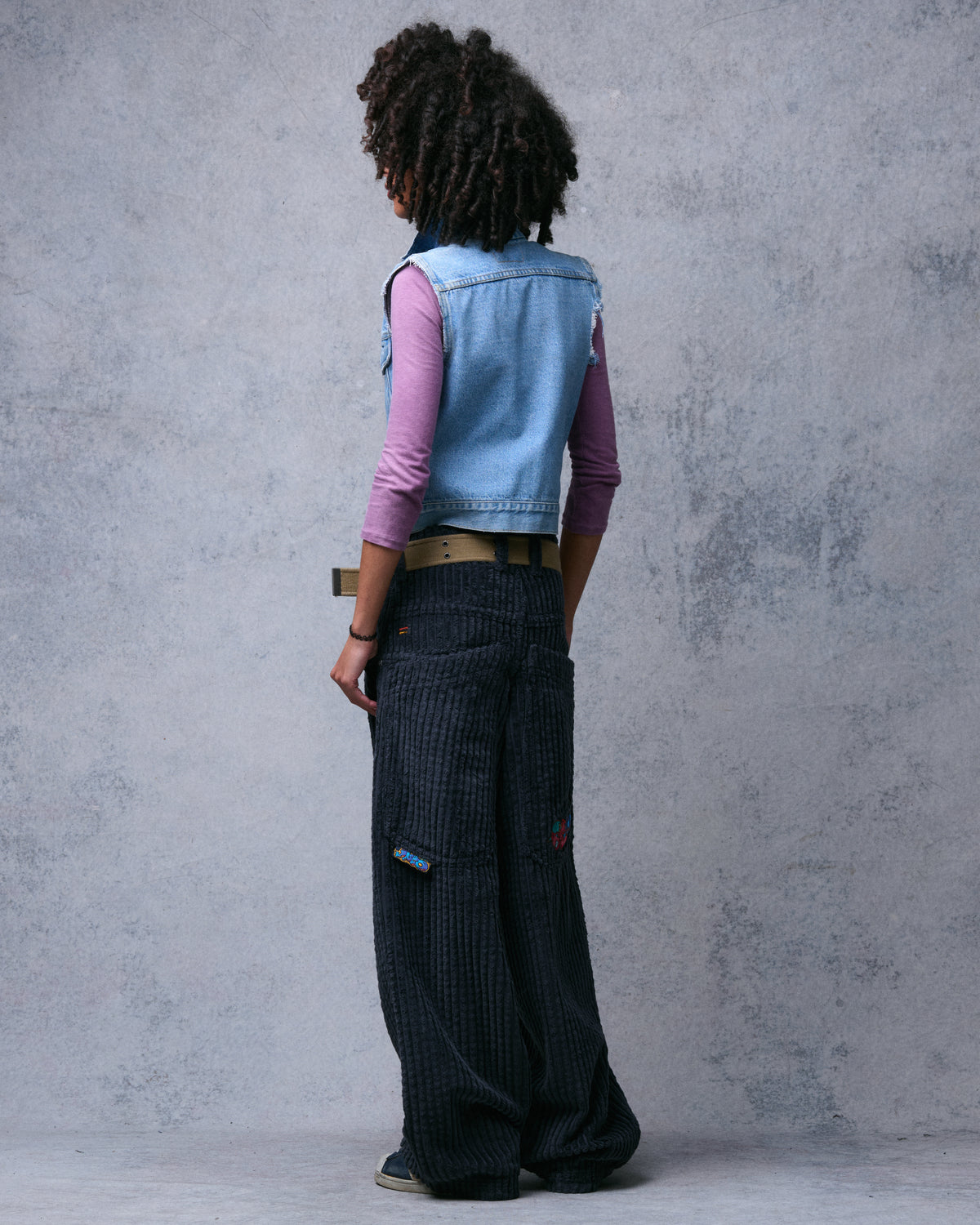 Model in dark charcoal wide-leg corduroy pants, angled back view showing the utility pocket tab and leg volume