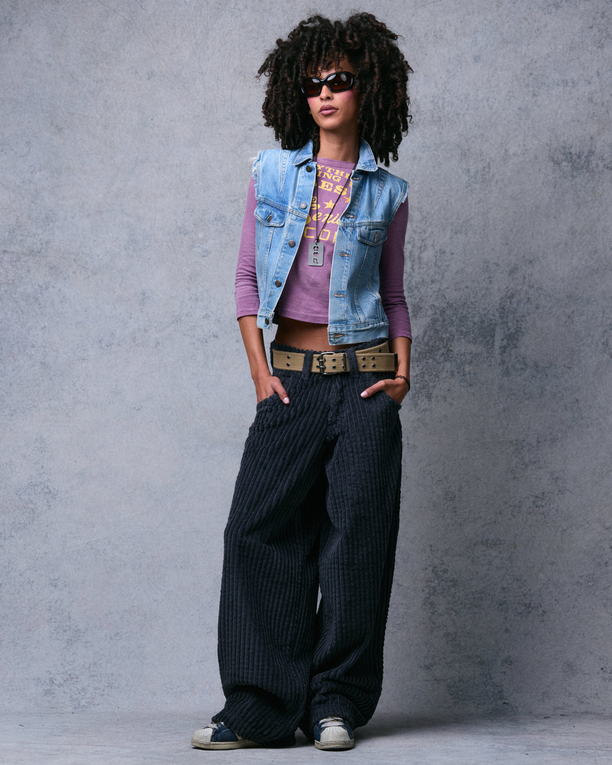 Model wearing dark charcoal wide-leg corduroy pants with a tan belt, cropped denim vest and sneakers, full-length front view