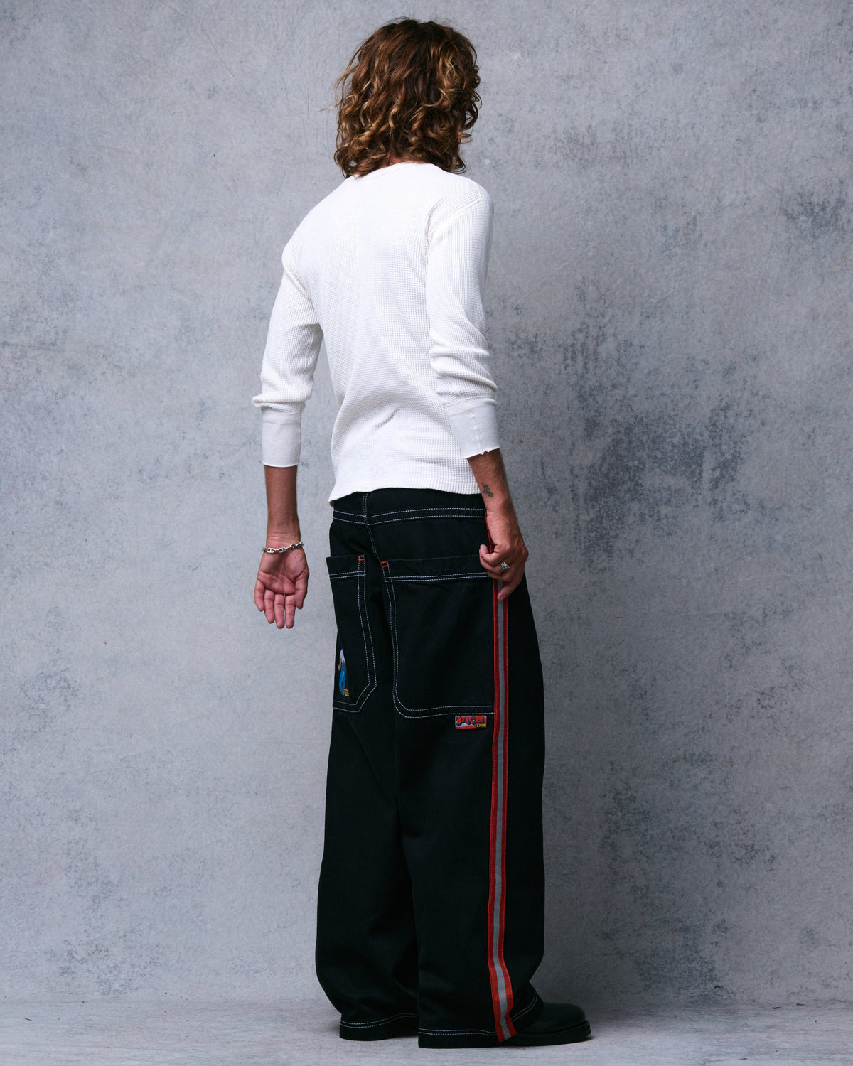 Model wearing wide-leg black jeans, angled back view showing the full-length orange and reflective side stripe
