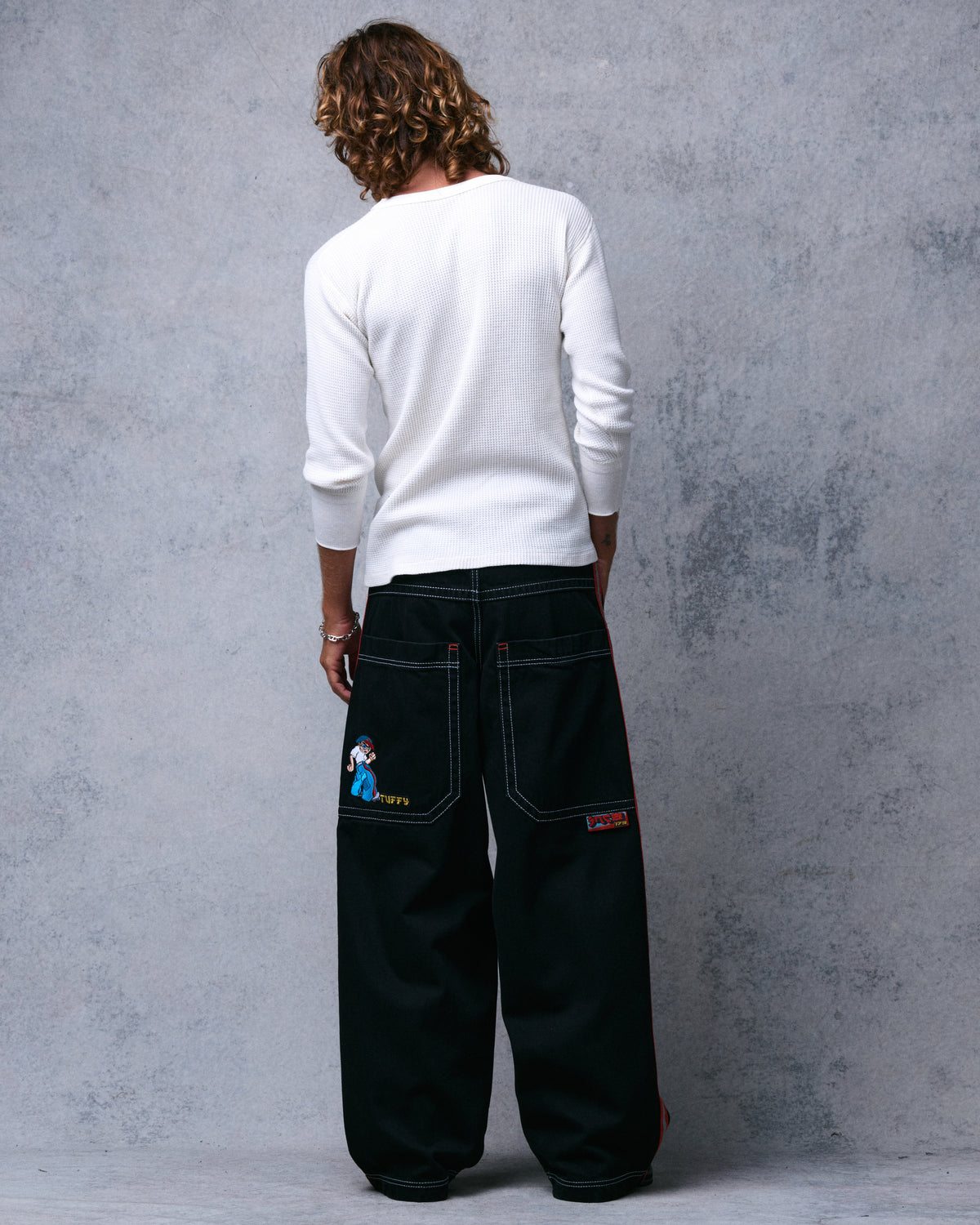 Model wearing wide-leg black jeans, straight-on back view of both pockets, embroidered character and patch