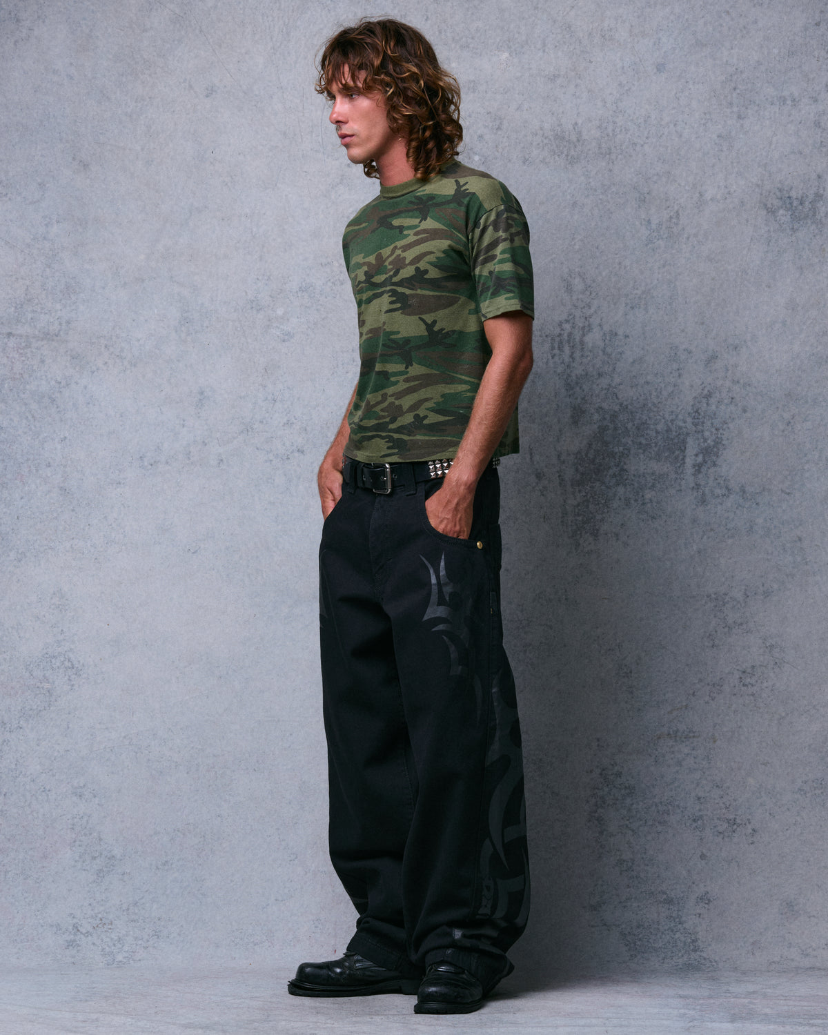 Model wearing wide-leg black jeans with tonal tribal print down the leg, studded belt and black boots, front three-quarter