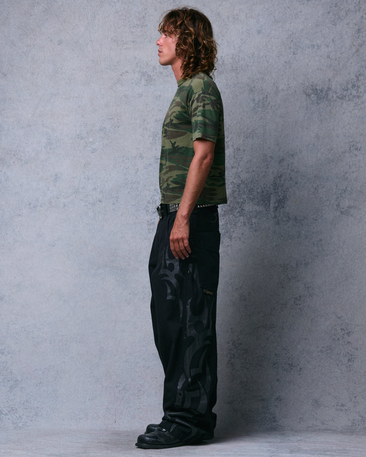 Model wearing wide-leg black jeans with tonal tribal print along the side and oversized back pocket, full side profile