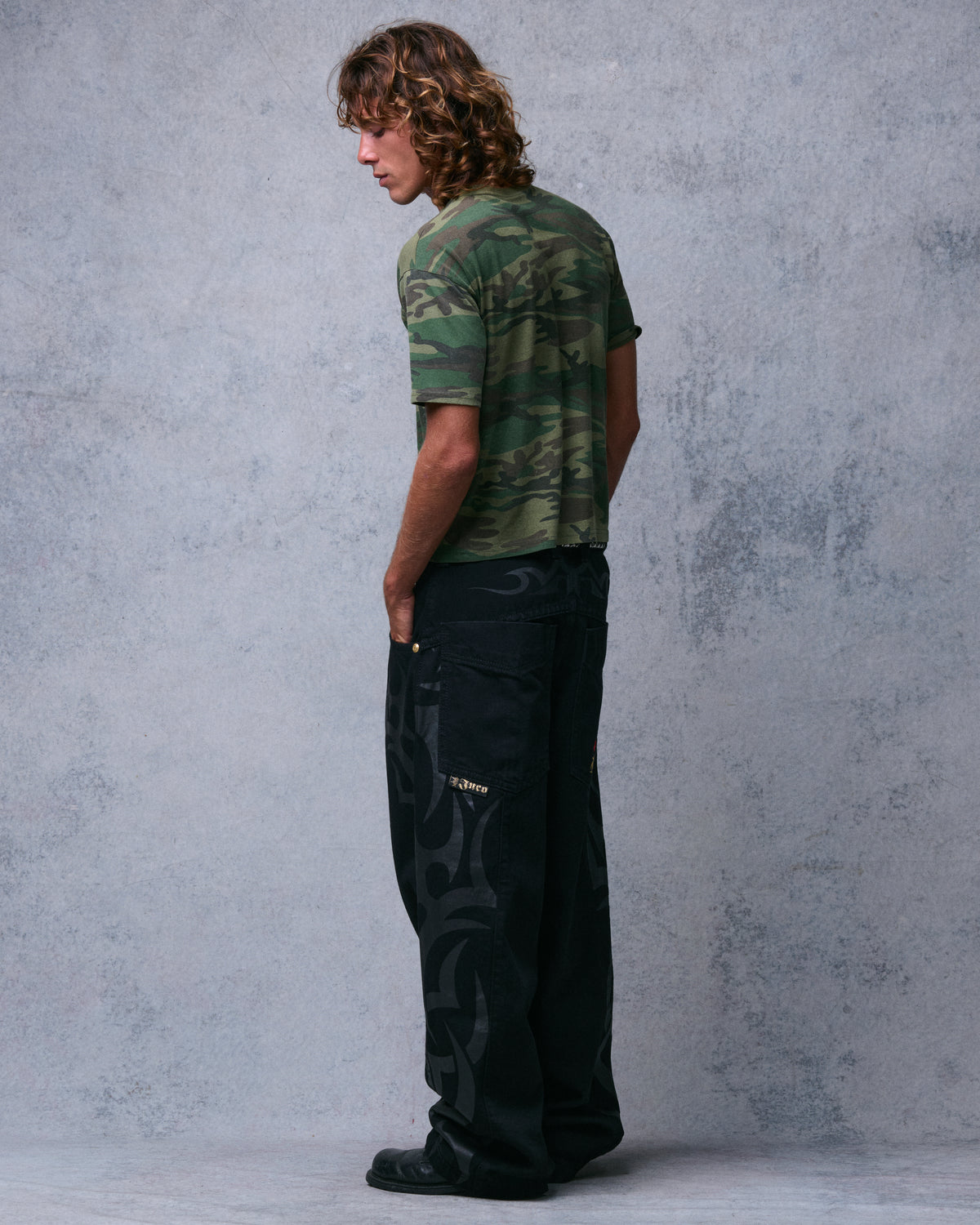 Model wearing wide-leg black jeans with tonal tribal print and oversized back pocket with Jnco tag, three-quarter back view