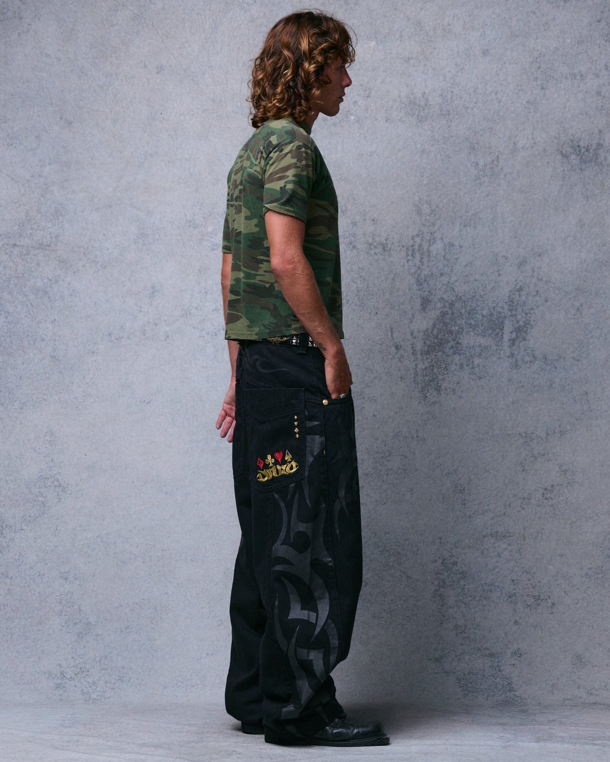 Model wearing wide-leg black jeans, side view showing tribal print down the leg and card-suit embroidered back pocket