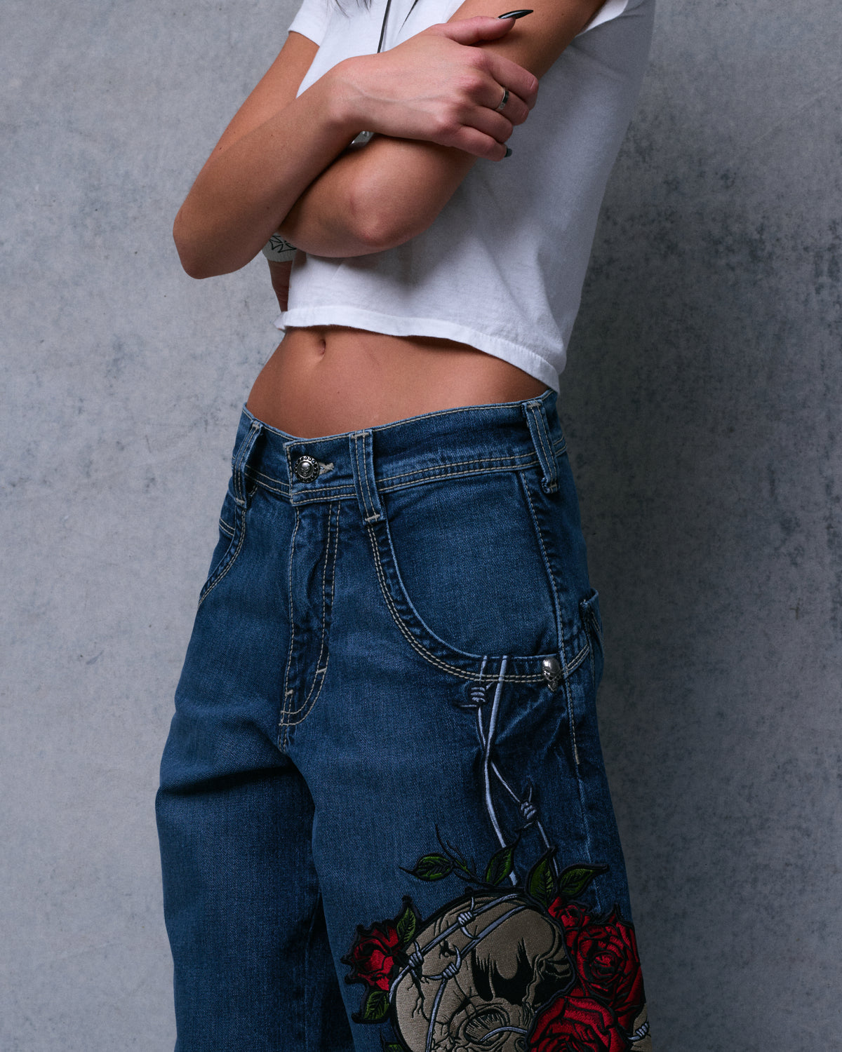 Model wearing the dark stone jeans, front three-quarter view of the waist with barbed wire and skull embroidery starting