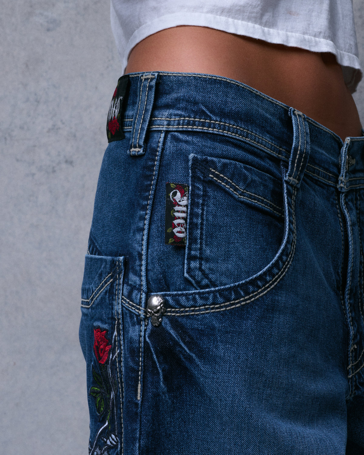 Side close-up of the jeans' hip showing a skull rivet, small embroidered rose patches and contrast stitching