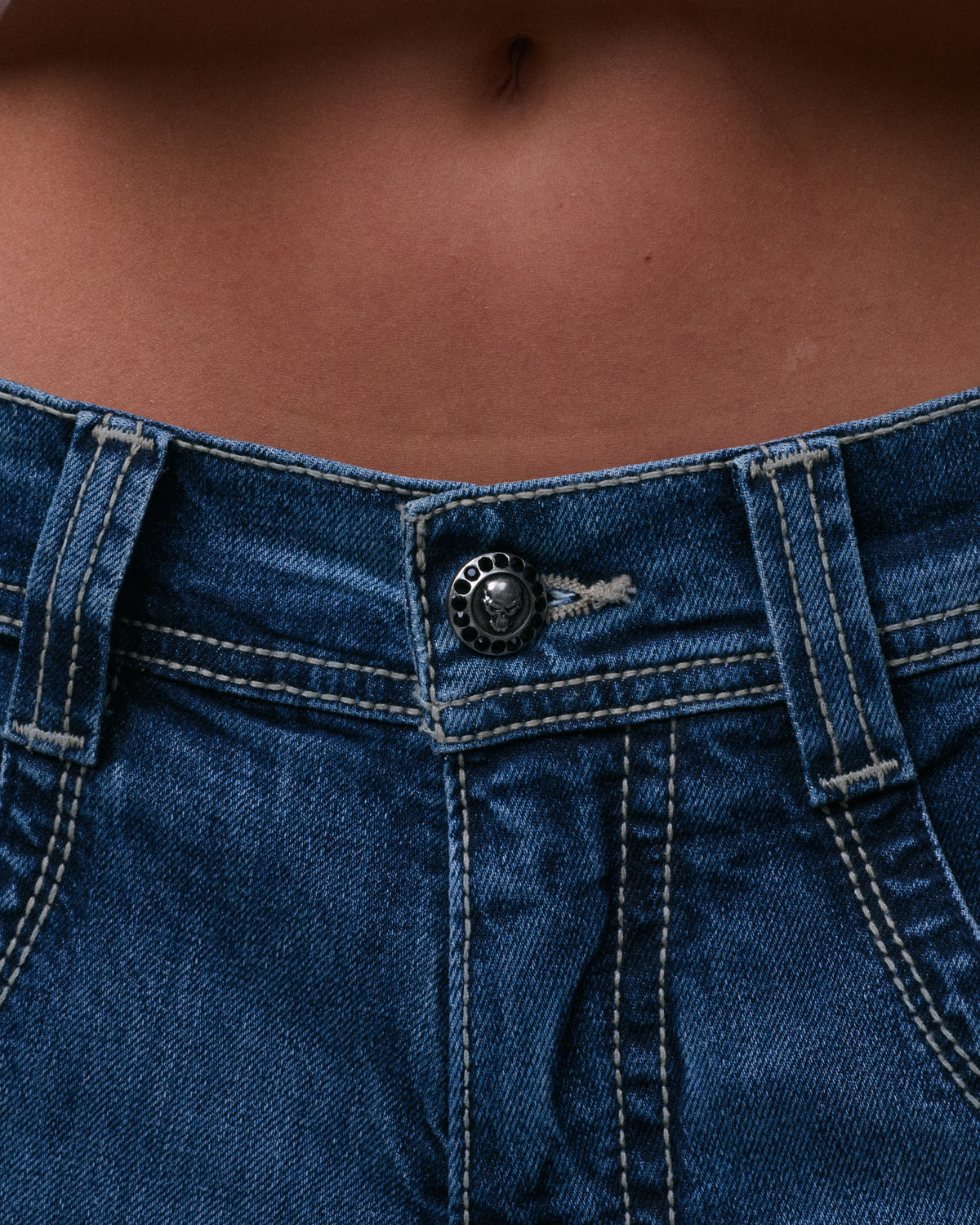 Close-up of the jeans' front waistband with a silver skull button ringed with black stones and contrast stitching