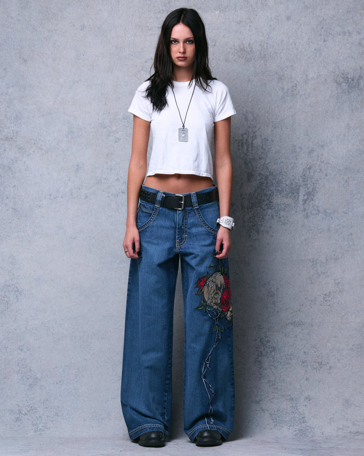 Model wearing dark stone wide-leg jeans with a belt, full-length front view showing skull and rose embroidery on one thigh