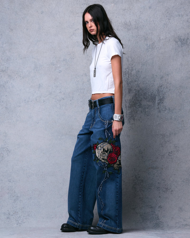 Model in the dark stone wide-leg jeans, full-length side view with the skull, roses and barbed wire embroidery facing camera