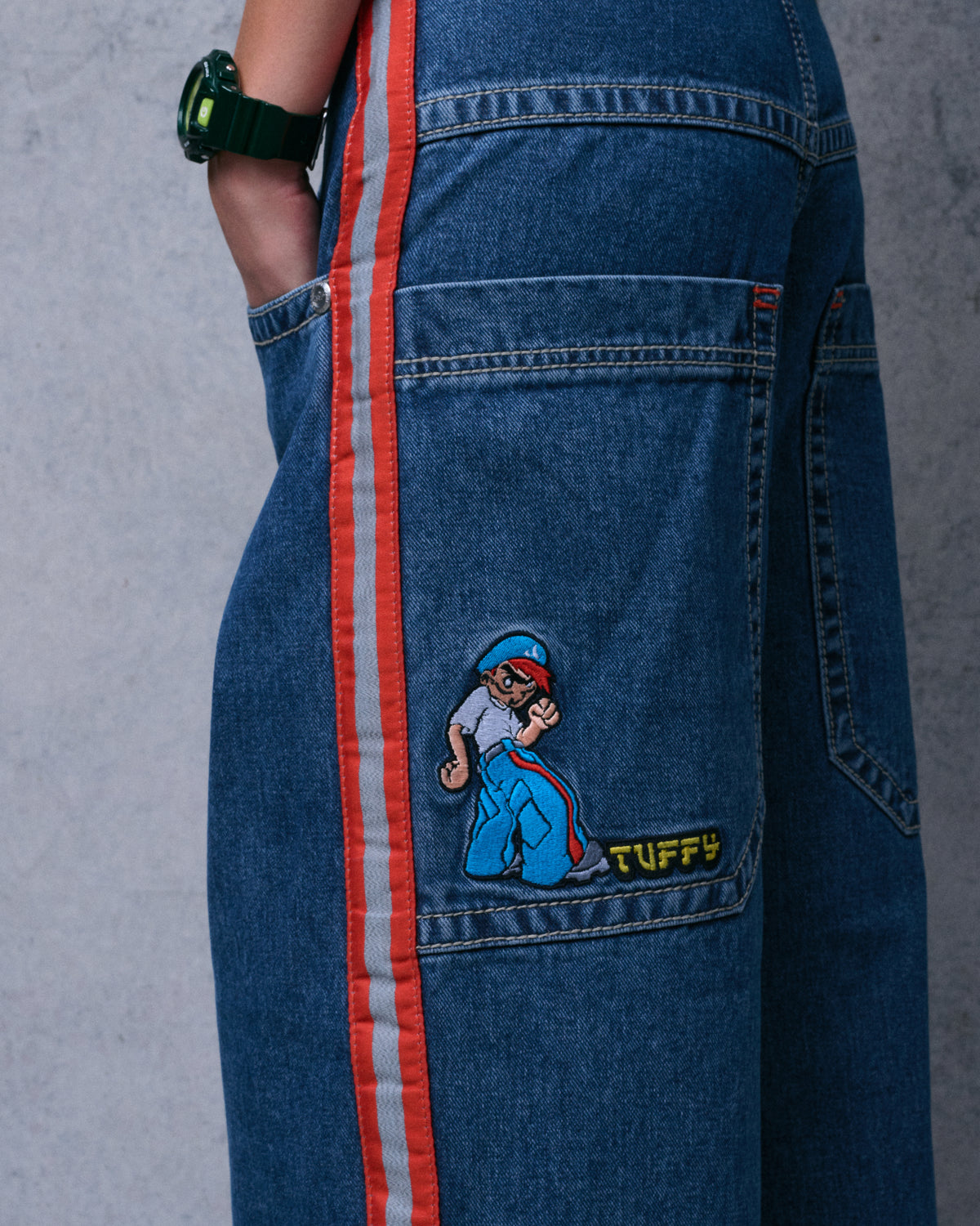 Side view of dark stone wash jeans showing an embroidered Tuffy cartoon patch on the back pocket beside the striped side seam
