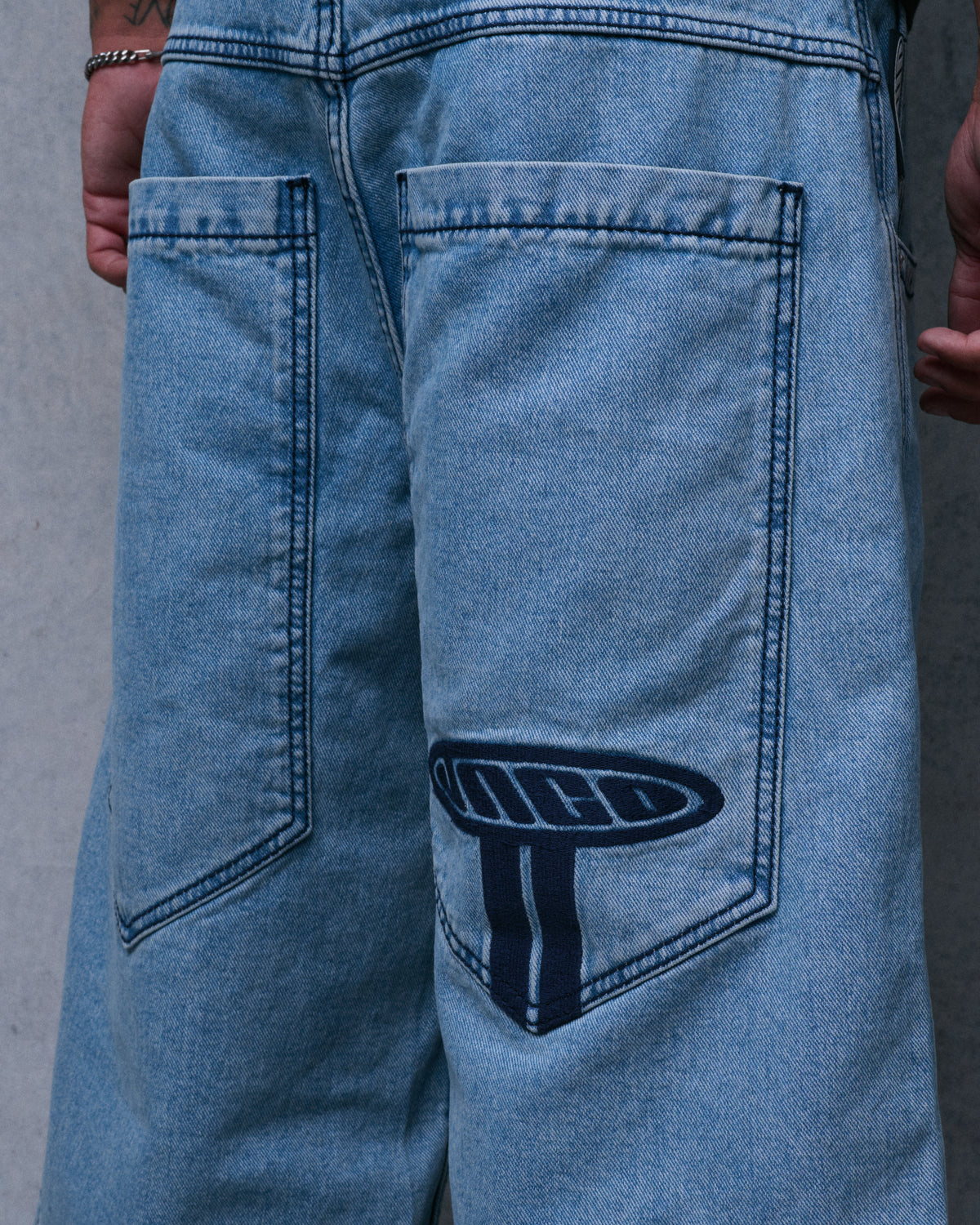 Close-up of the elongated back pockets on light blue denim shorts with navy JNCO embroidery on one pocket