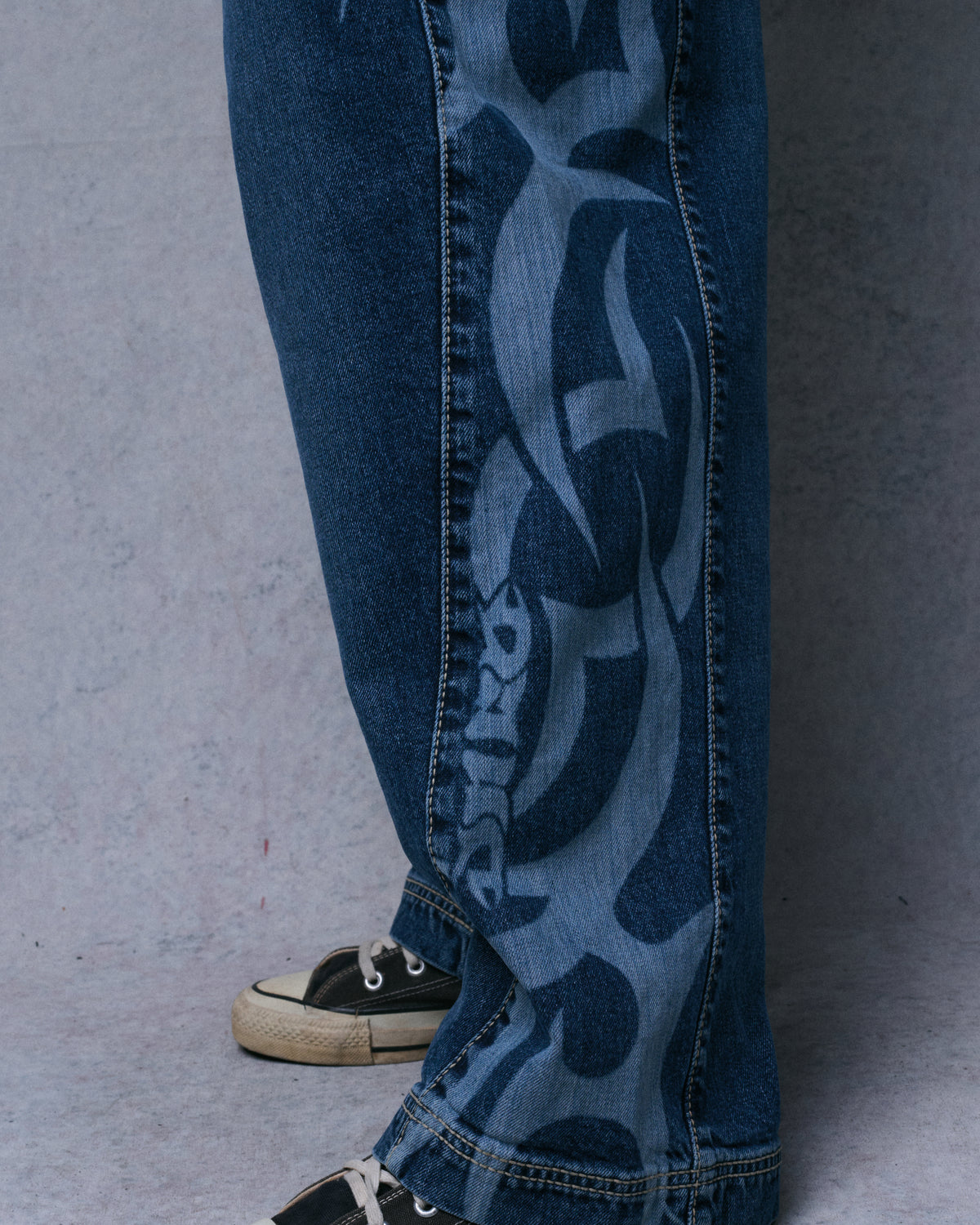 Close-up of a wide jeans leg with laser-etched tribal graphics down the side seam, worn with canvas sneakers