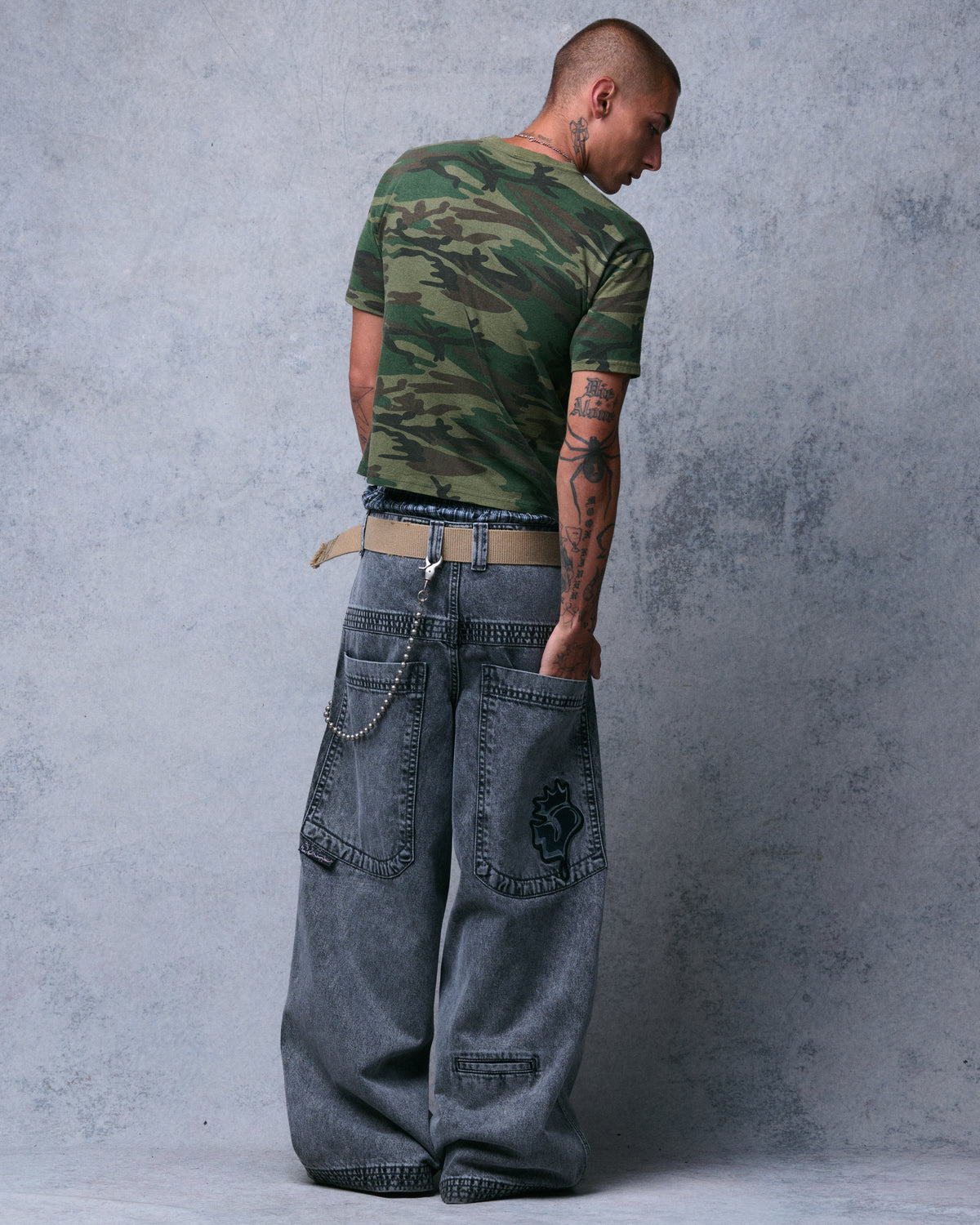 Model wearing faded black wide-leg jeans with a tan belt and wallet chain, back view, one hand in the Crowned J back pocket