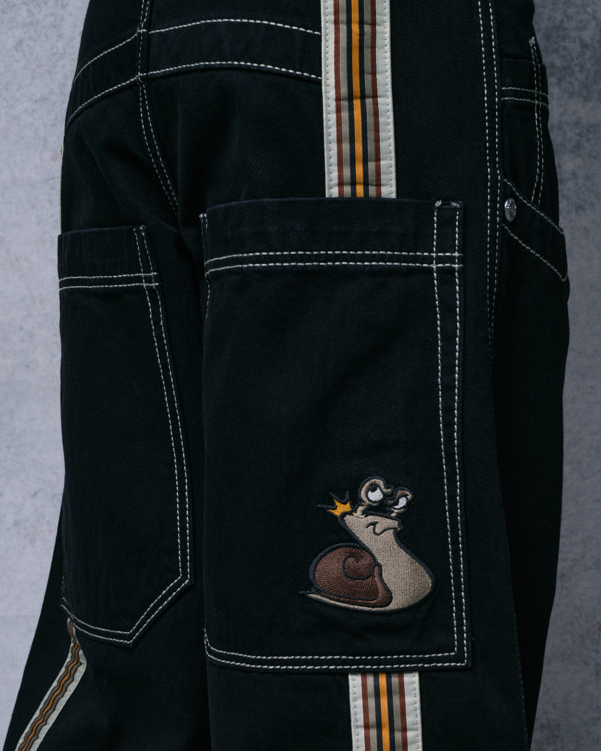 Close-up of a black jeans back pocket with an embroidered snail patch wearing a crown, beside the striped tape