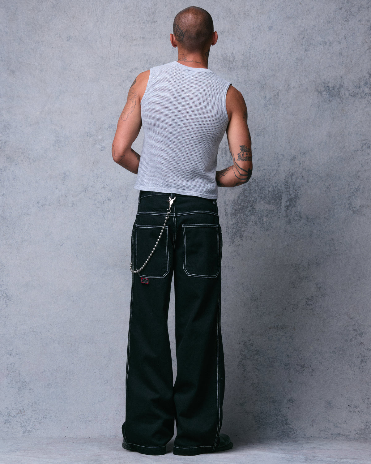 Model wearing wide-leg black jeans with white-stitched back pockets, red logo tab and wallet chain, back view
