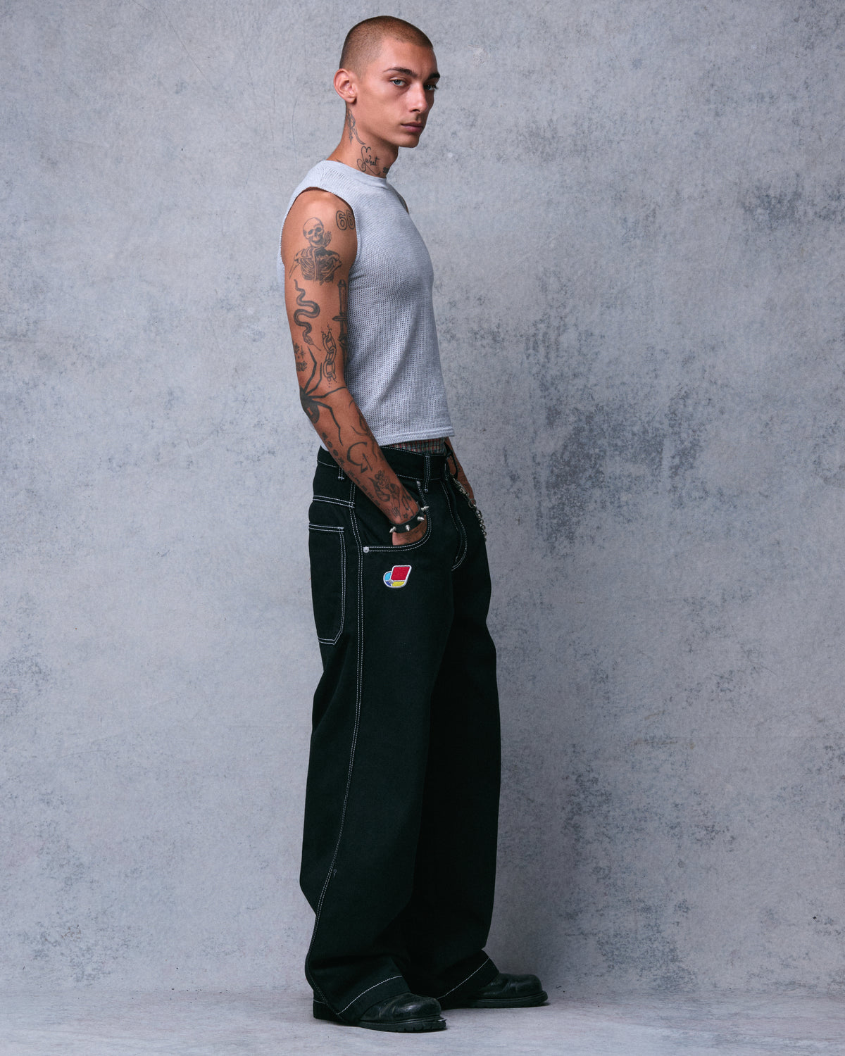Model wearing very wide-leg black jeans with white stitching and an embroidered pocket patch, side view