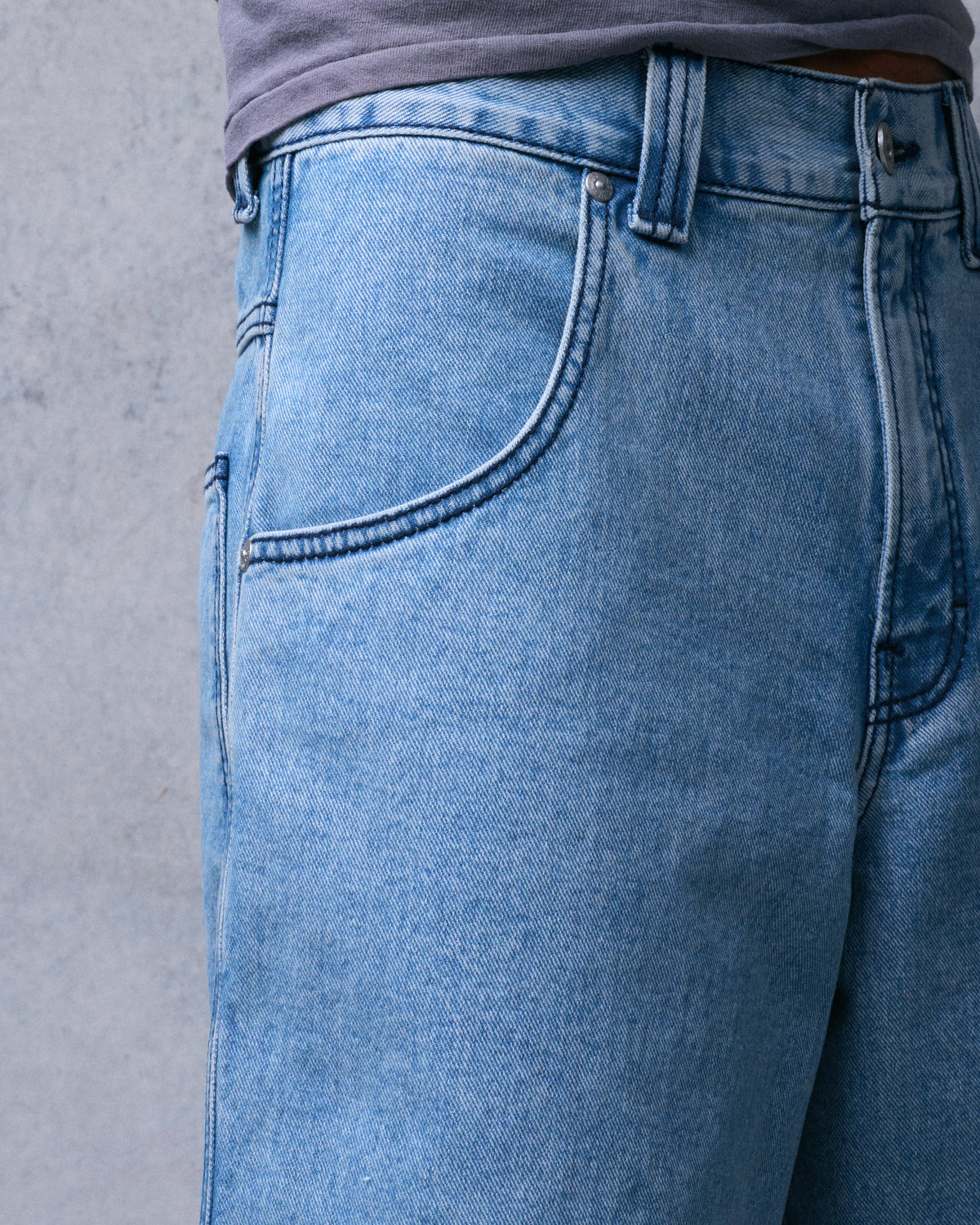 Side-front close-up of light blue denim shorts showing the front pocket, rivets and button waistband